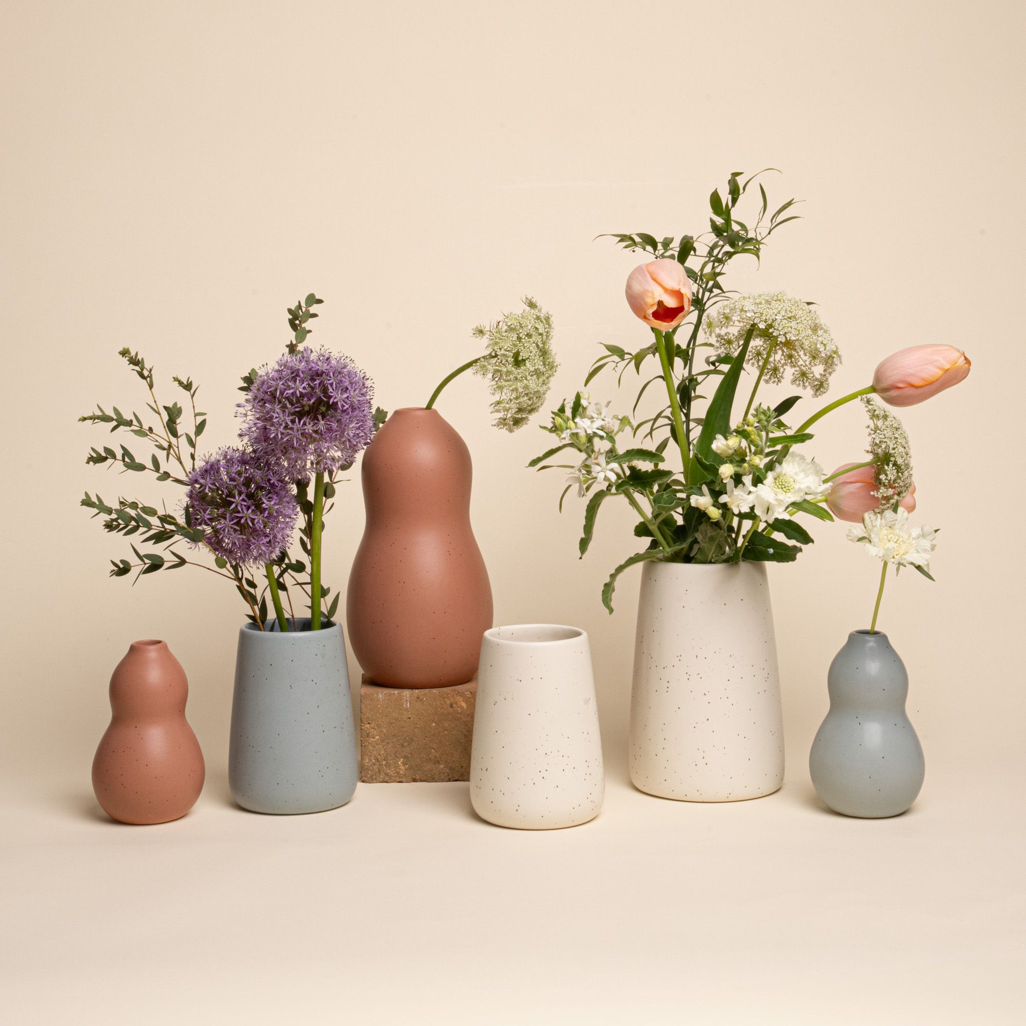 A lineup of two different sized and three different colored vases wth fresh blooms in them. One vase has a double gourd and the other is a more classic slope-style vase. They come in three colors: Utah, Pebble and Panna Cotta.