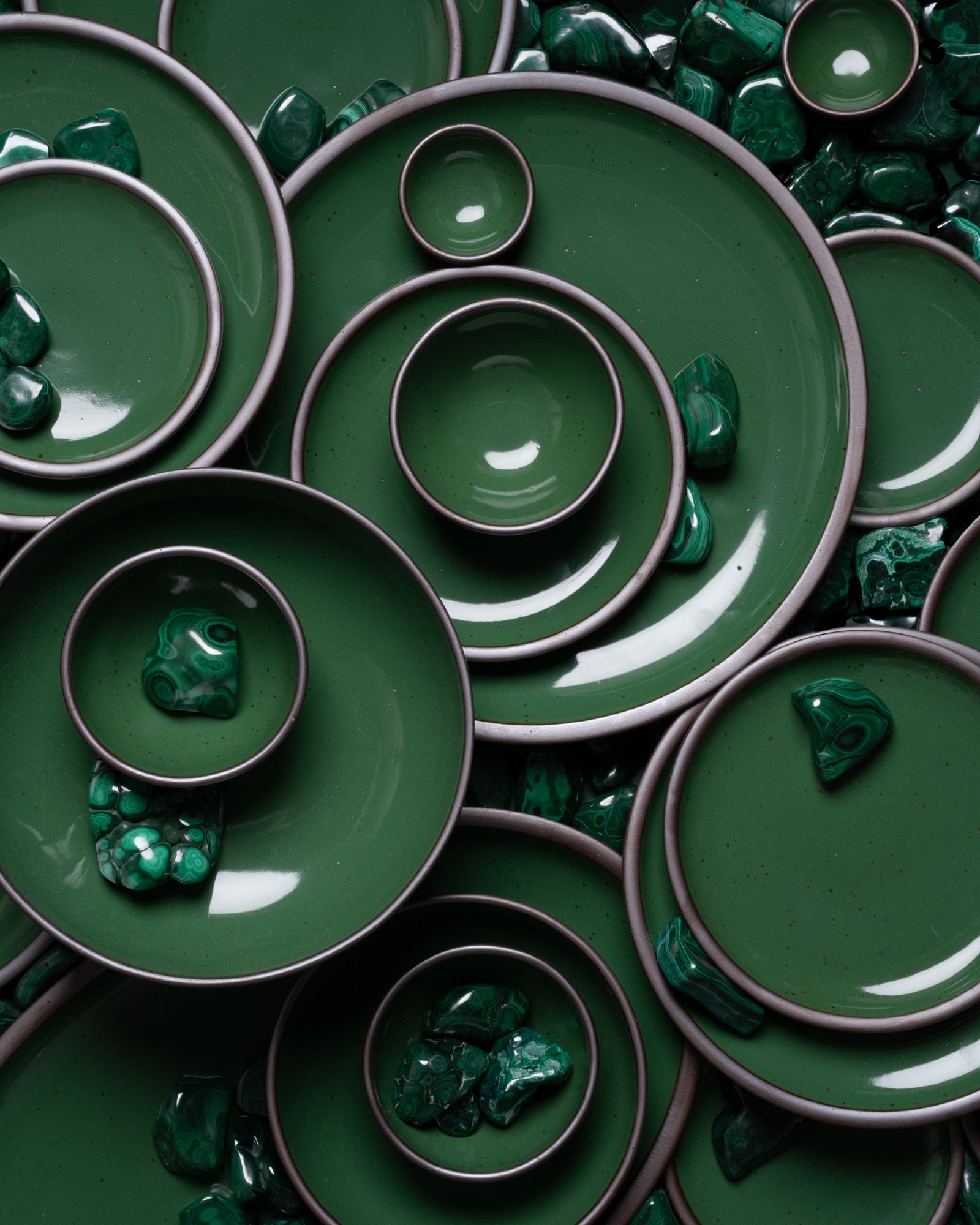 Arrangement of glossy emerald green ceramic plates and bowls with scattered malachite stones on a dark green background.