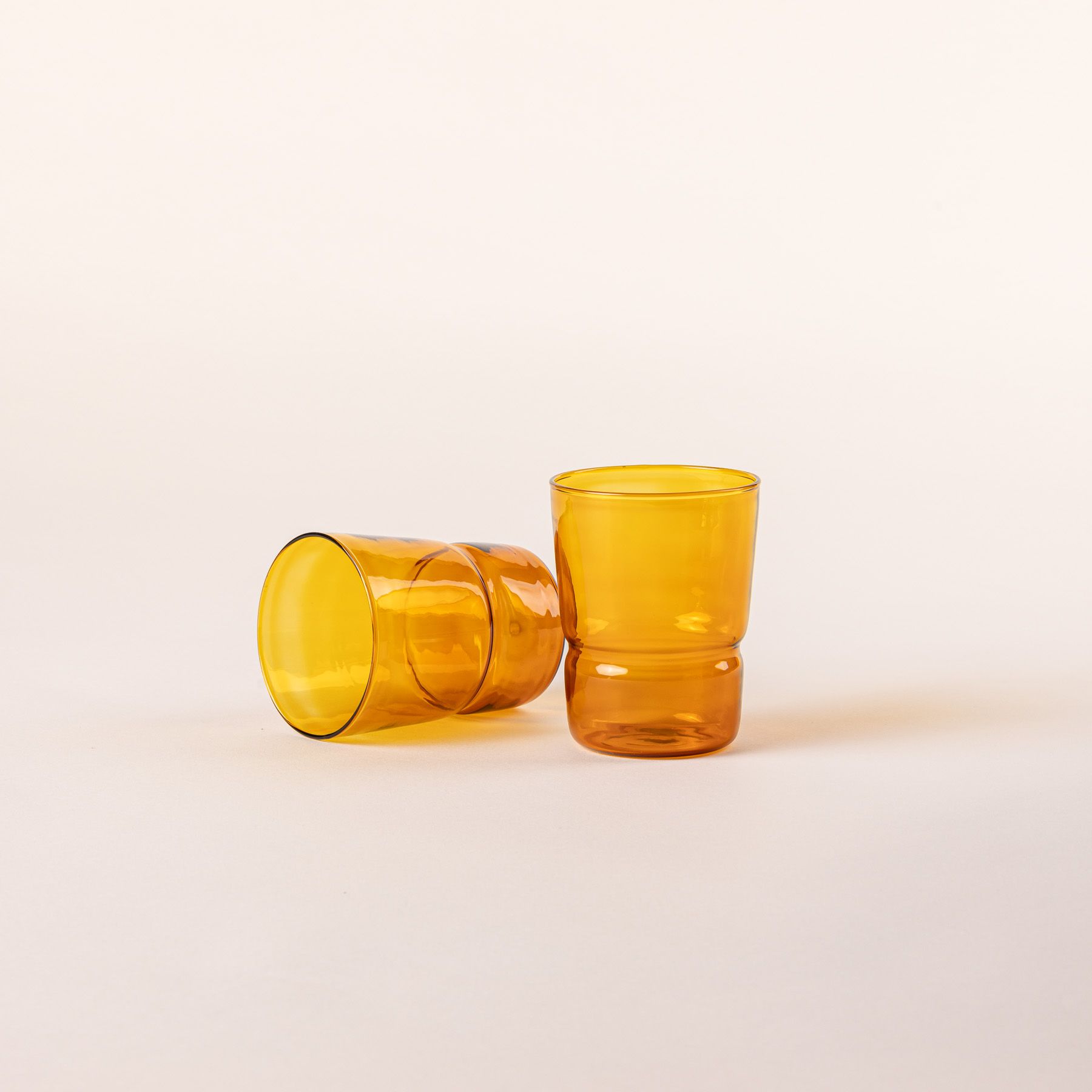 Two tumbler glasses that widens at the top in an amber color, 1 is standing up and 1 is laying on the side.