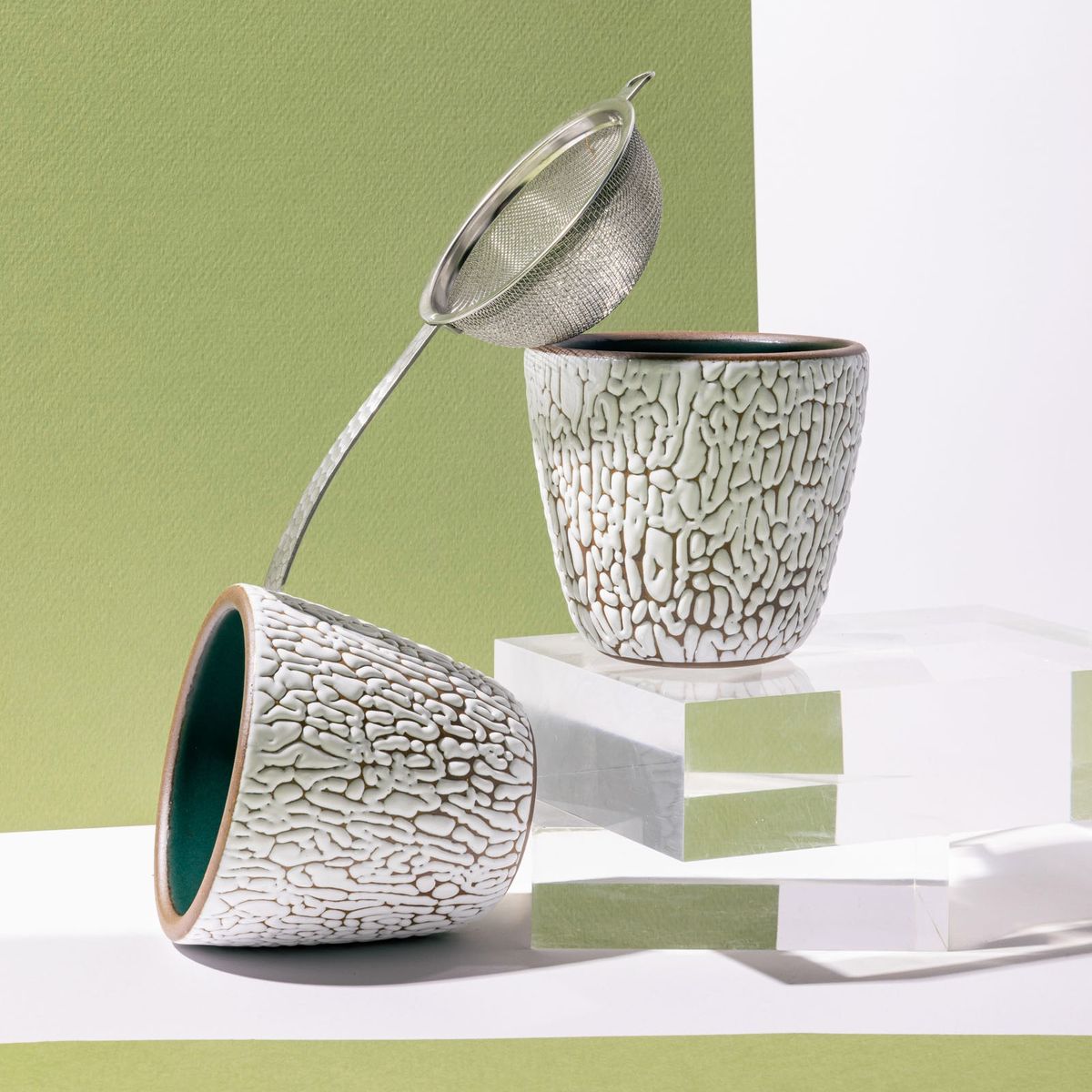 Two small ceramic cups with cracked textures and the interior being dark teal and arranged with a steel strainer