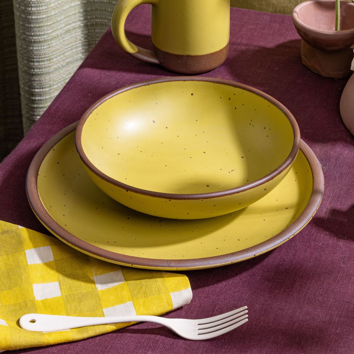 A dinner-sized shallow bowl sits on a dinner plate, both in zesty yellow colors, on a deep purple tablecloth with an enamel fork and more.