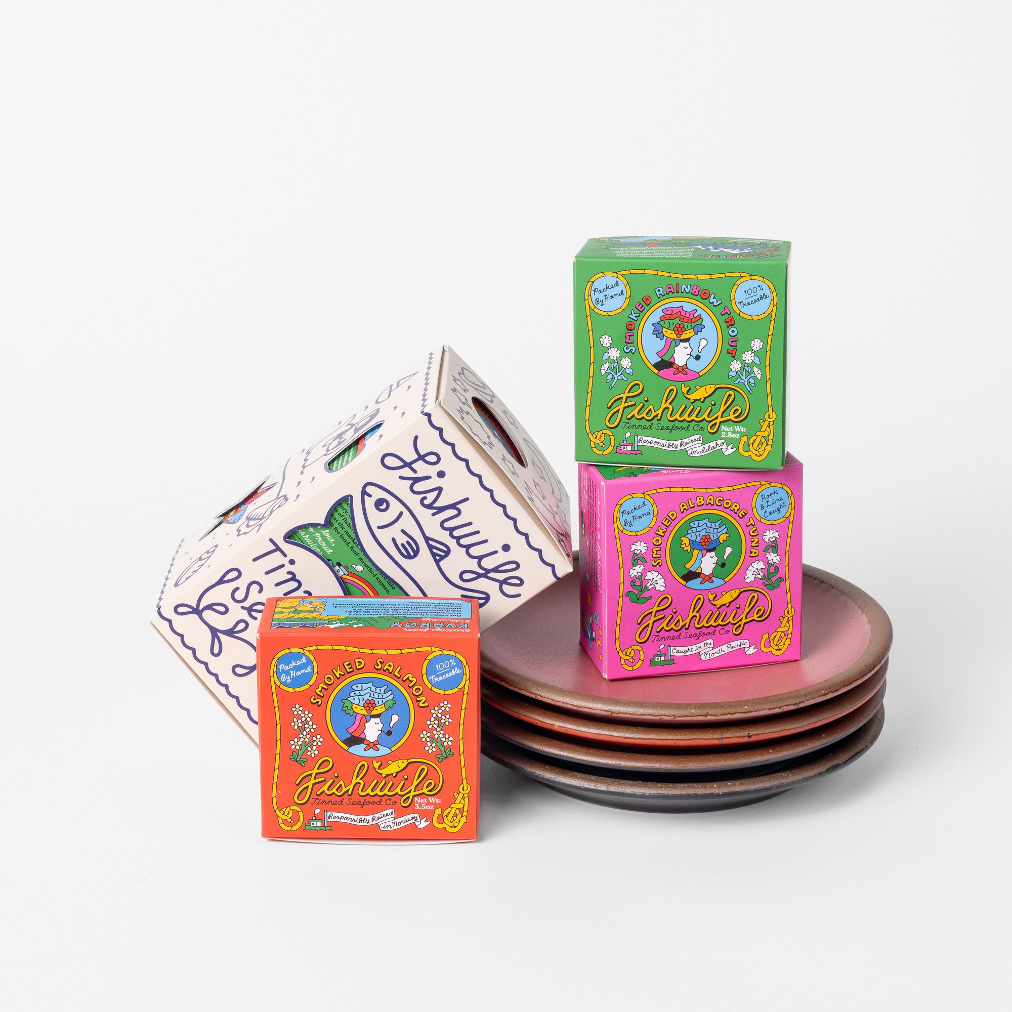 A stack of four ceramic plates with a stacked square boxes of tinned dish displayed on top. The boxes are illustrated in white, green, pink, and orange colors.