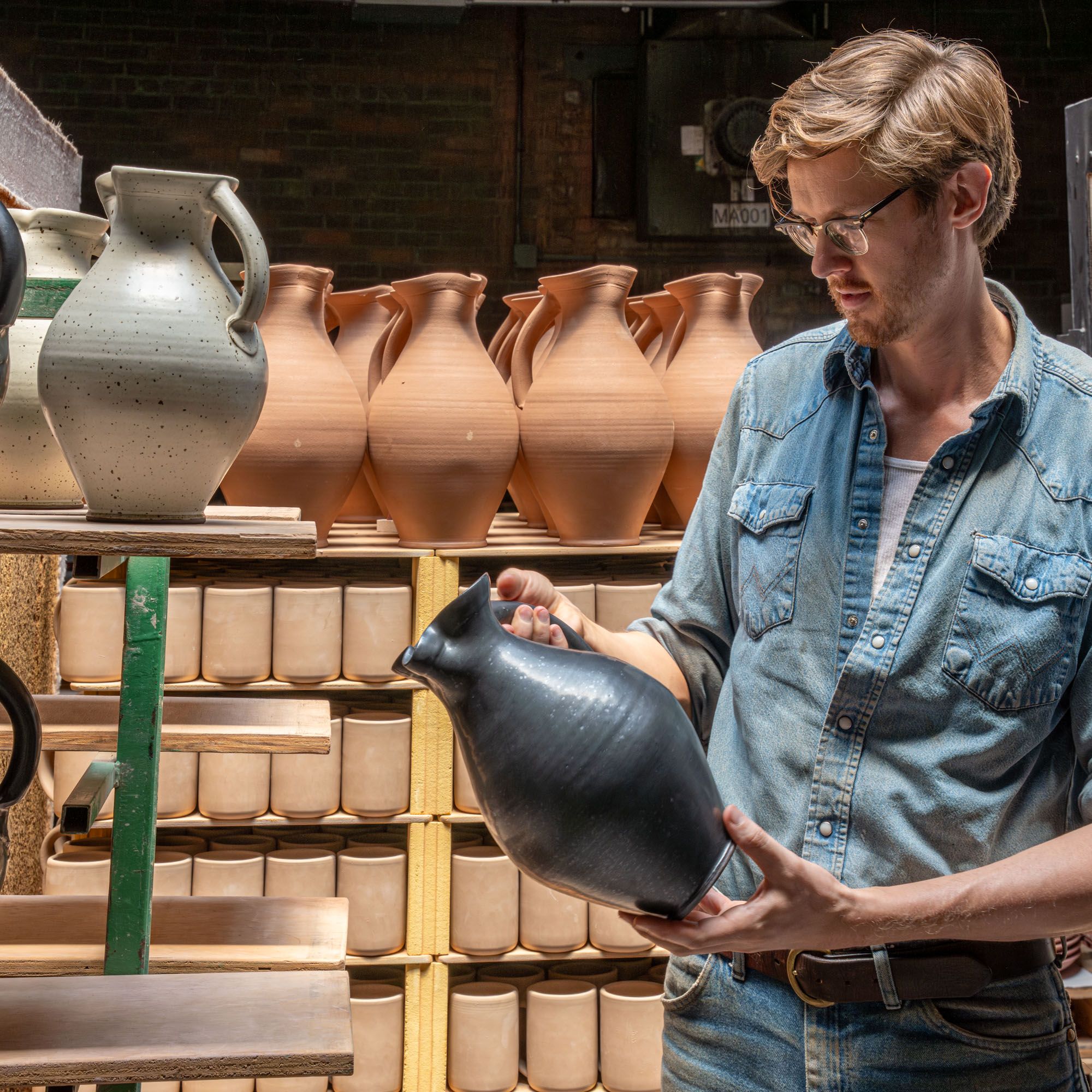 John holding a finished pitcher in the factory in front of pitchers ready to be fired in the kiln.