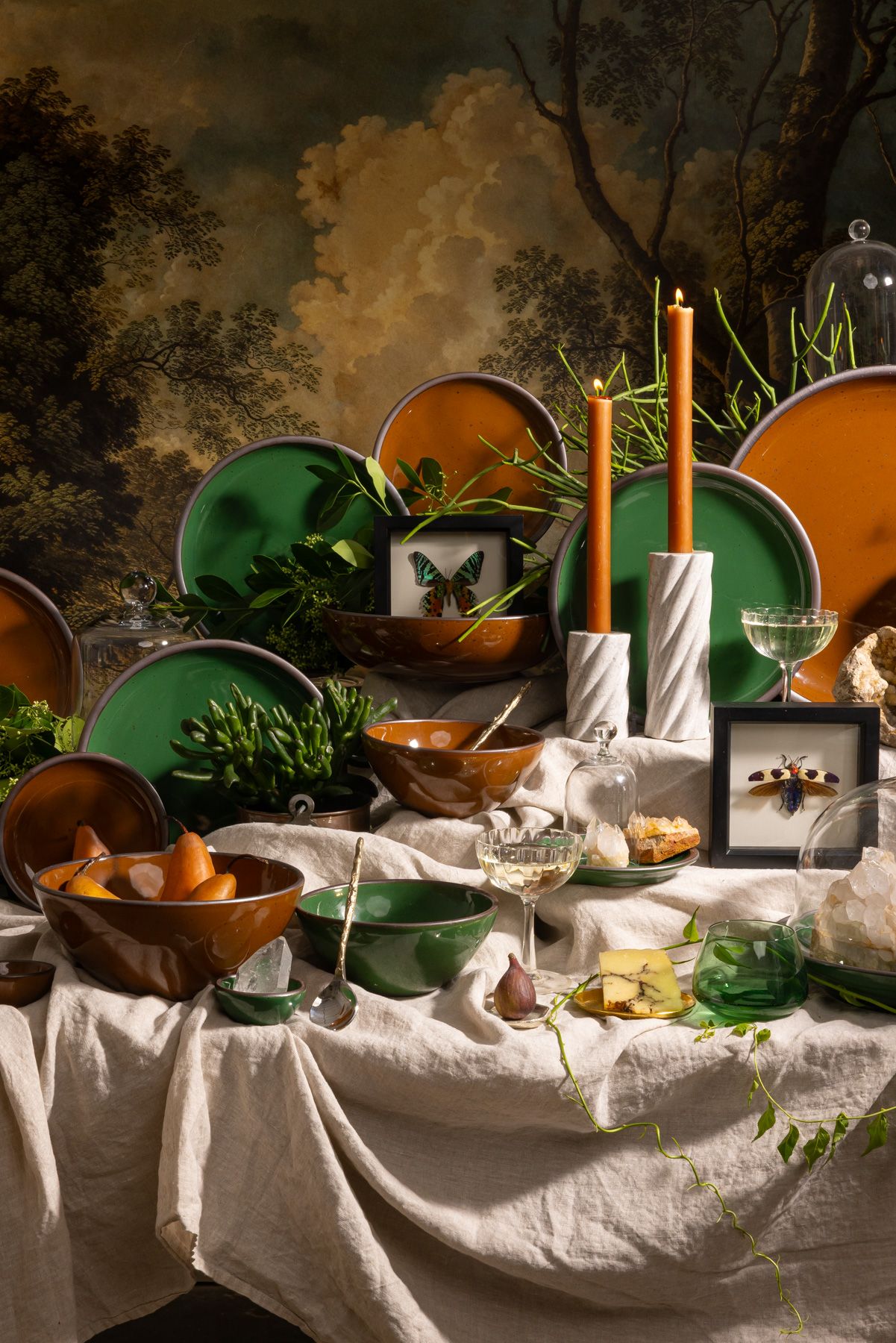 Table with glossy green and brown ceramic plates, candles, crystals, plants, and framed insects against a forest mural backdrop.