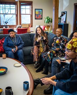 A diverse group of people are gathered inside on a sectional couch in conversation around a table
