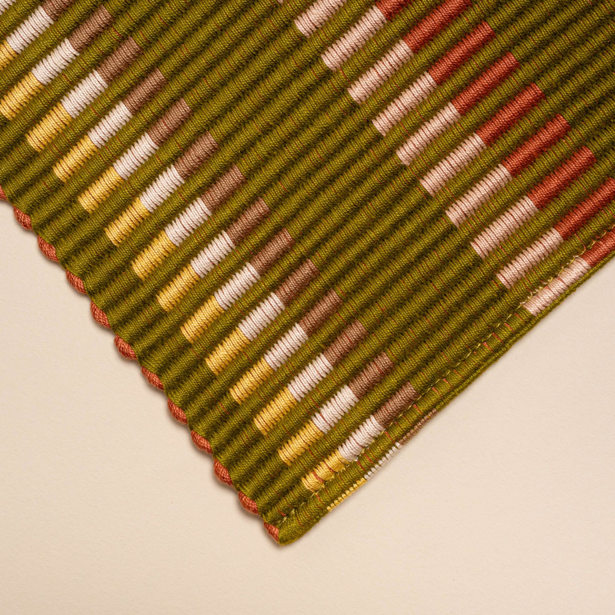 A corner of a rectangle placemat with offset stripe design that is hand woven in olive, tan, orange, and yellow colors