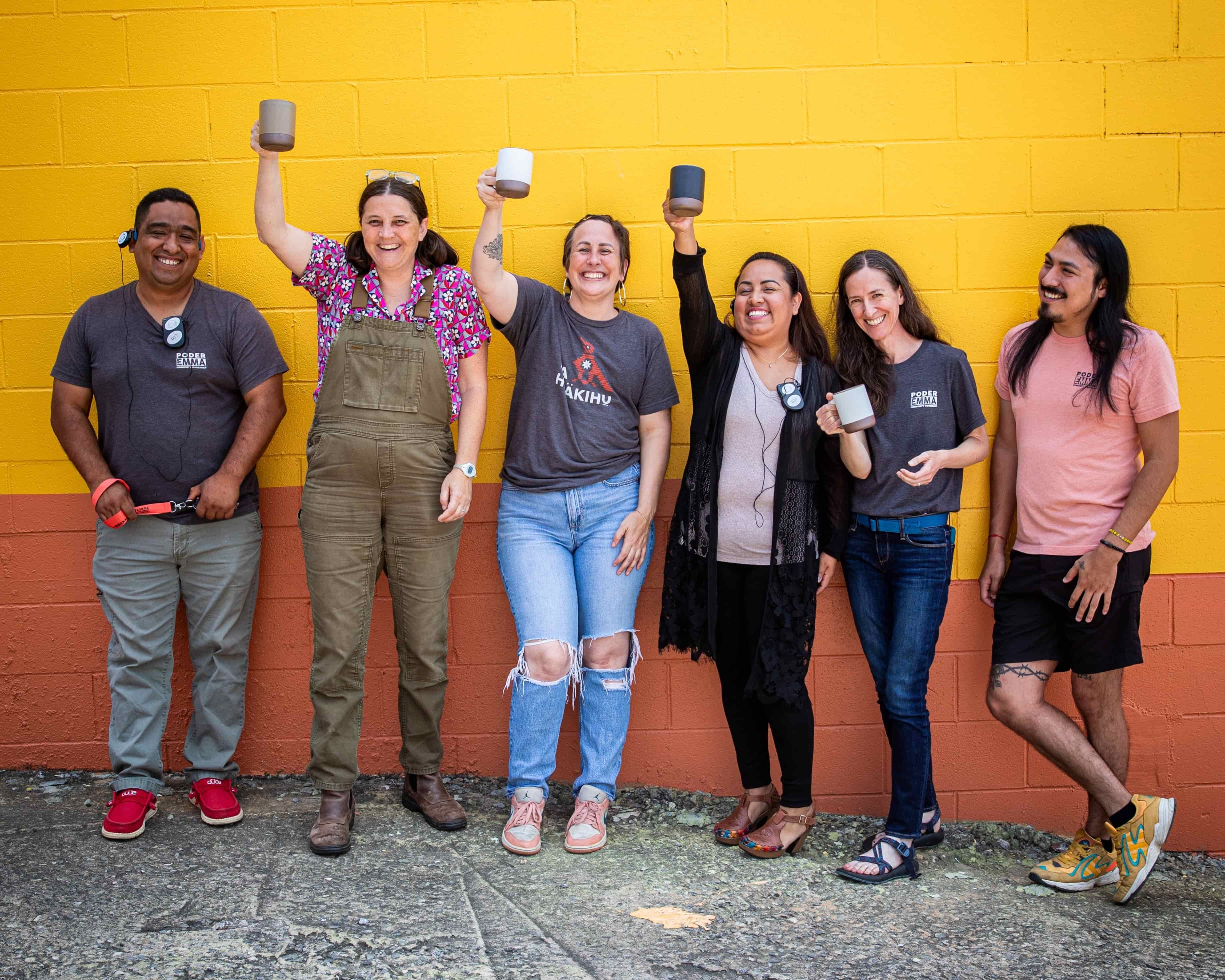 A group of people against a yellow wall outside, smiling and holding East Fork mugs in the air.