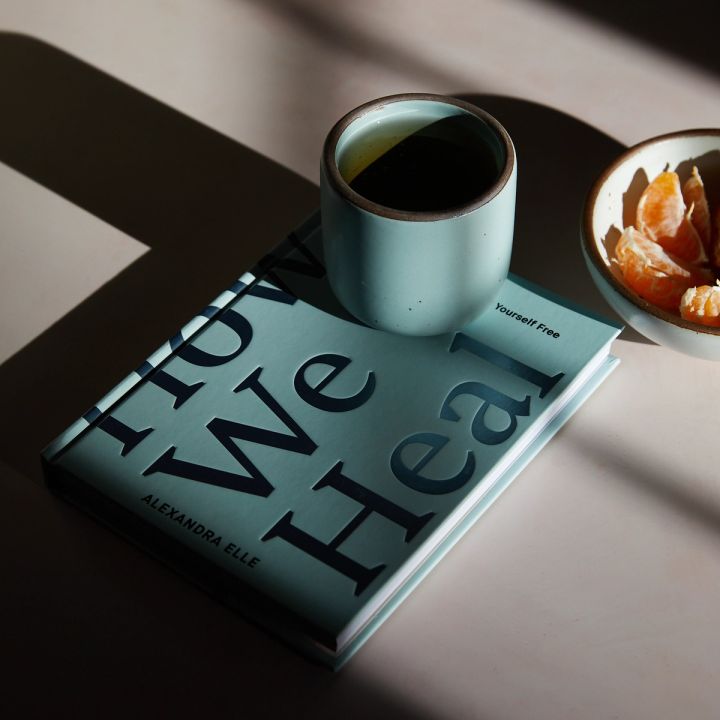 A book that reads 'How We Heal' with a ceramic cup in Malibu sitting beside it with shadows. A little bowl of orange slices is close by.