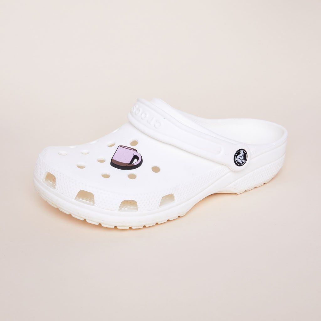 A white plastic clog made by the brand Crocs with a pale purple charm on the top that looks like an East Fork mug