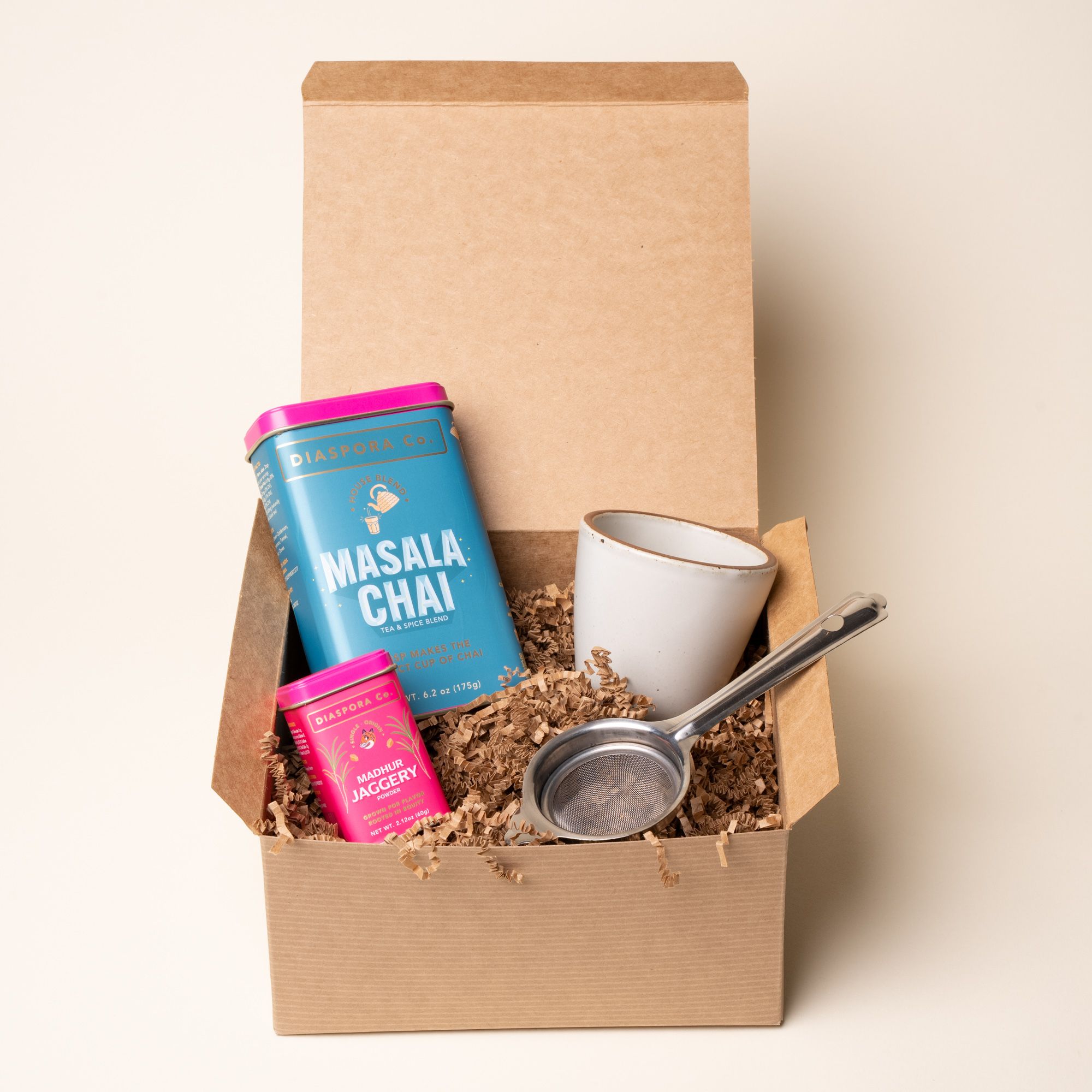 Gift box with Diaspora Co. masala chai blend, jaggery, a ceramic cup, and a metal strainer nestled in kraft paper filling.