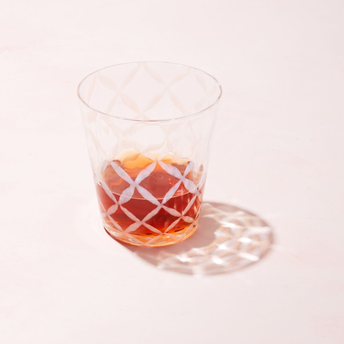 A short clear glass with a delicate grid pattern, half filled with drink and casting a shadow