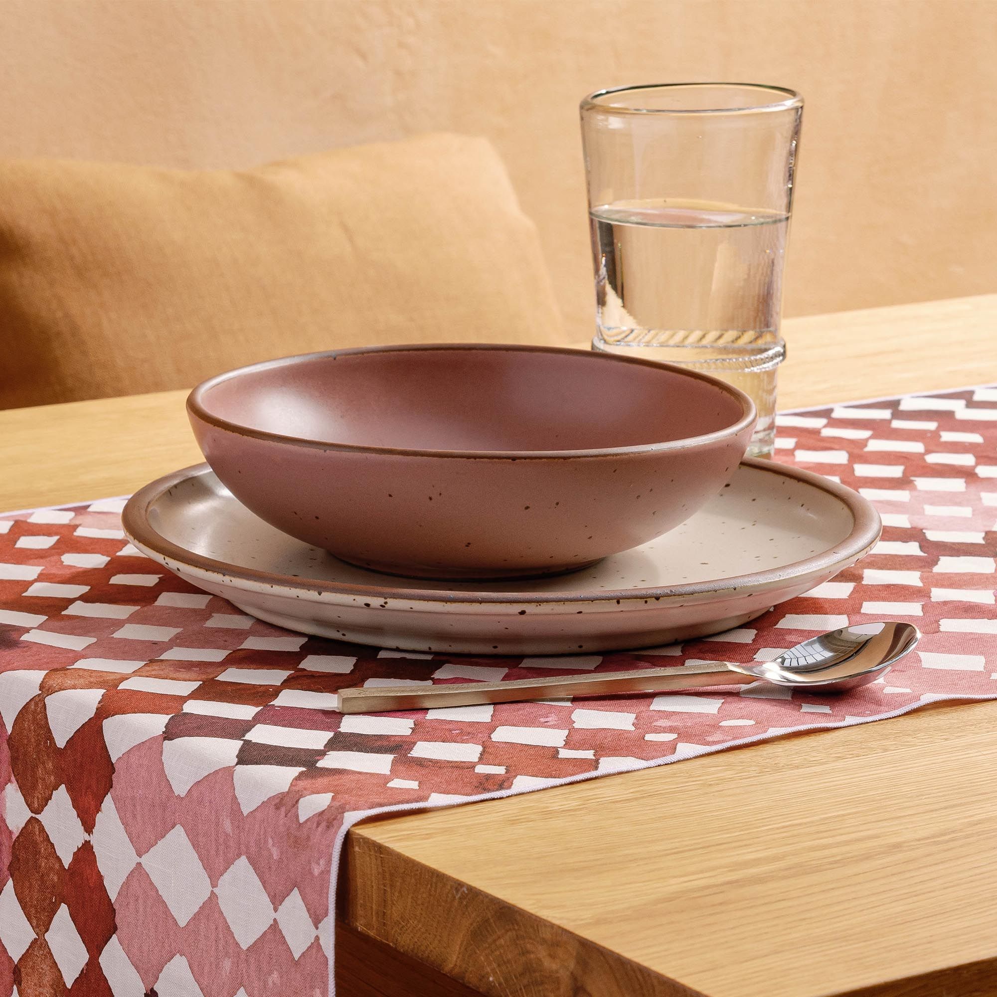 Dusty mauve pink ceramic shallow bowl on a plate with a spoon and glass of water on a patterned table runner.
