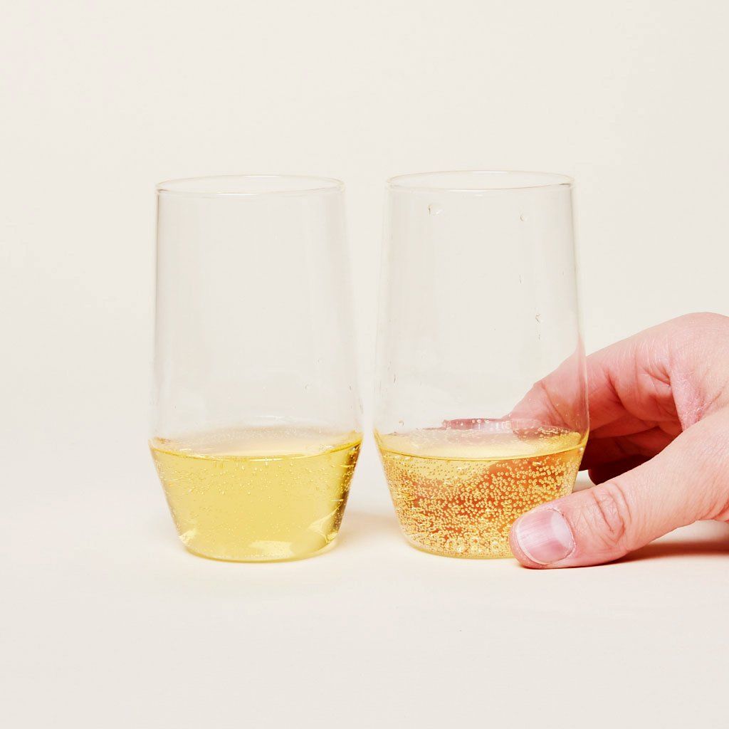 A hand rests on one of two tall stemless wine glasses that taper toward the base, filled about one-third with bubbly, yellow liquid