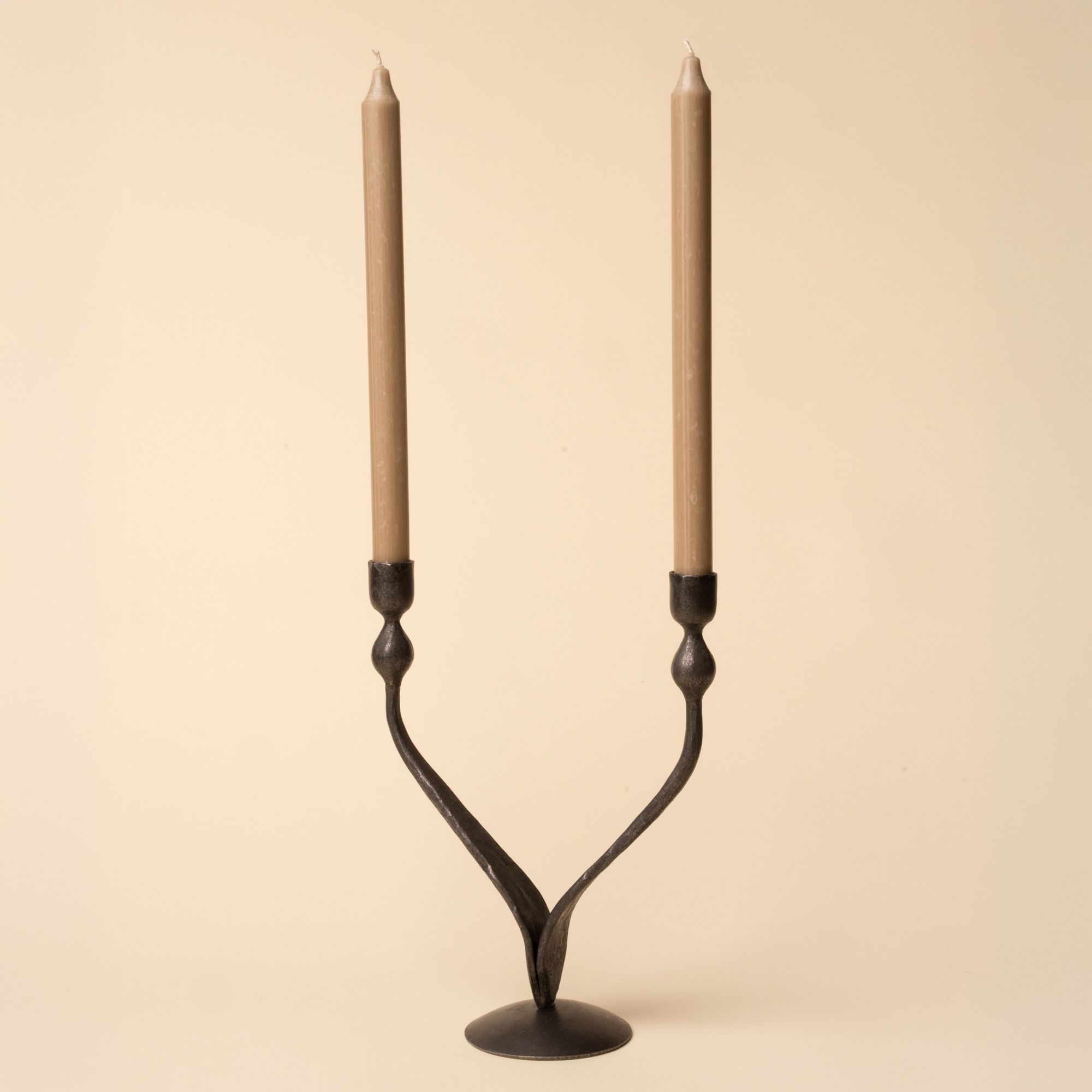 Forged iron two-armed candelabra with a Y-shaped design, holding two tall taupe taper candles, displayed against a neutral beige background.