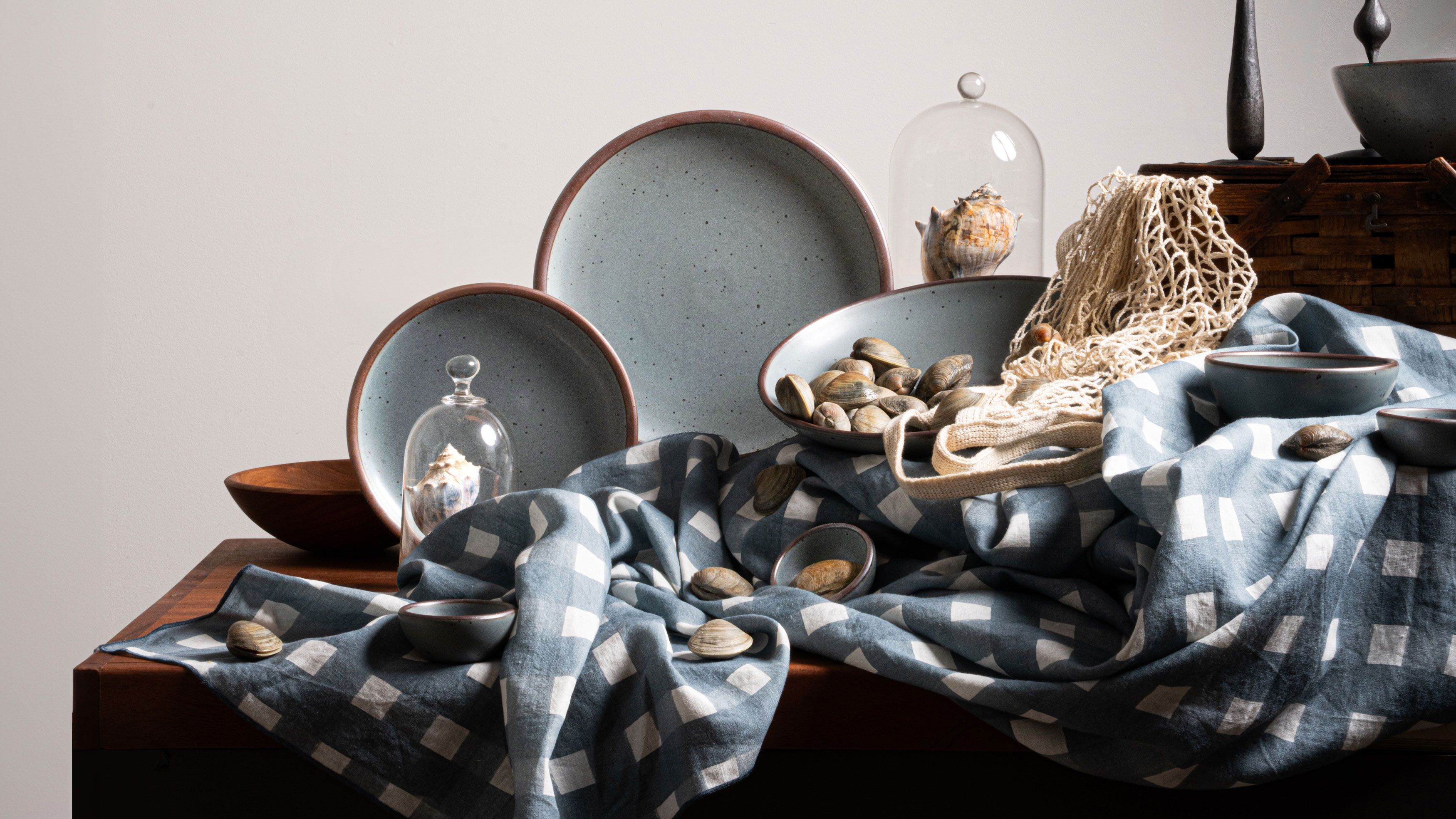 Ceramic plates in a blue-grey color artfully arranged on a table with blue-grey crosshatch fabric, shells, bell jars, and a taper candle.