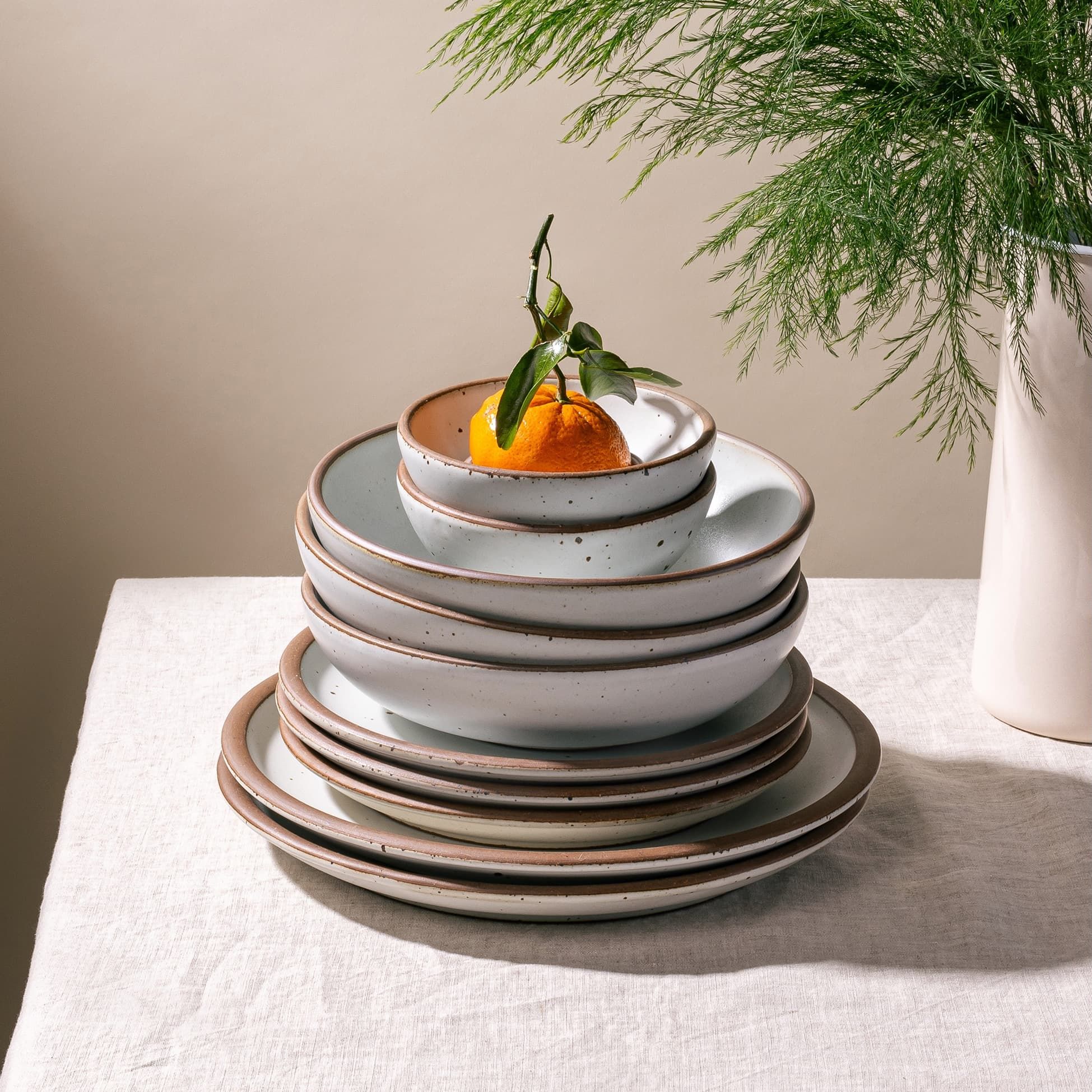 On a table sits a stack of ceramic bowls and plates in cream and cool white colors with an orange on top. To the right is a cream pitcher with greenery coming out of the top.