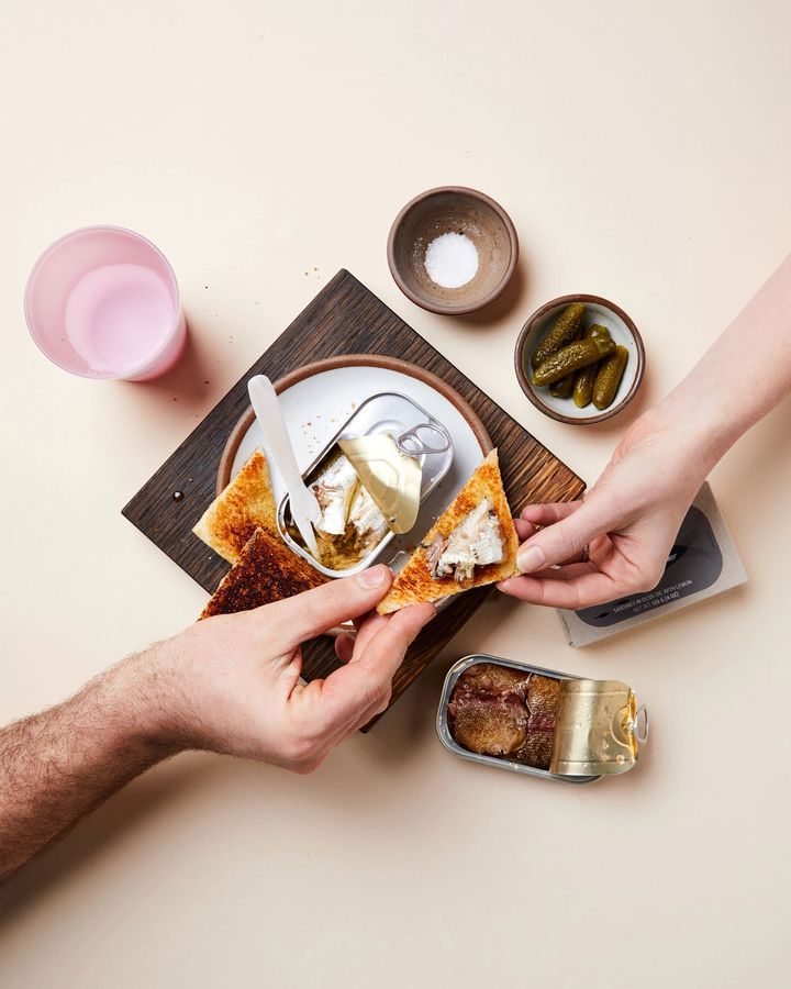 Wooden cutting board topped with plate, tinned fish and toast. Two hands reach in for the toast.