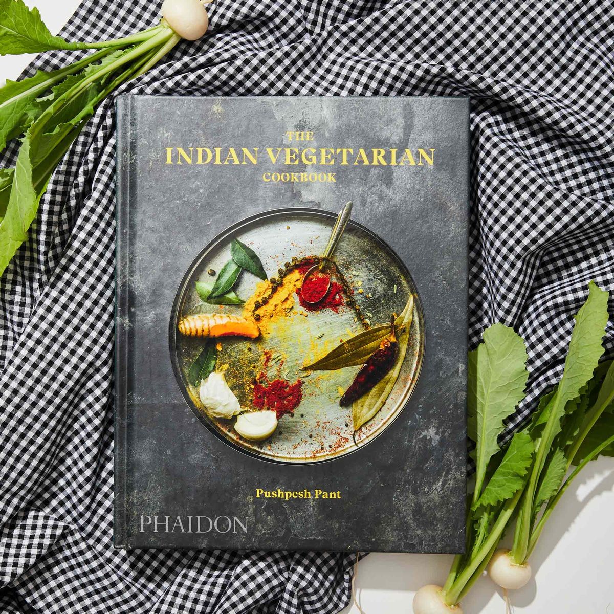 The Indian Vegetarian by Pushpesh Pant resting on a piece of black and white gingham fabric, with radishes around it