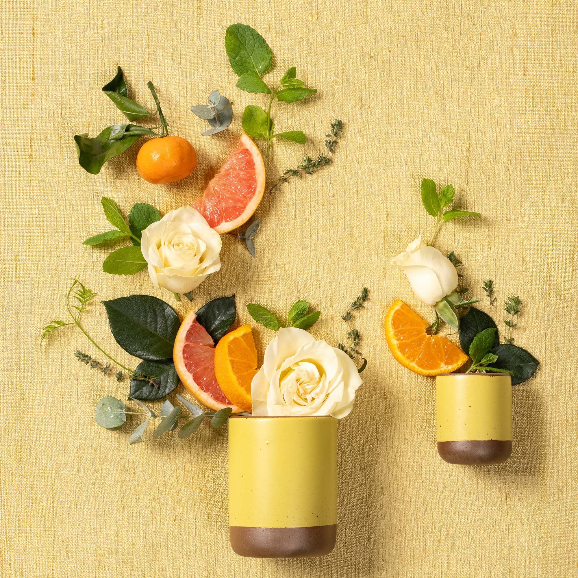 A small and large cylindrical ceramic vessels in a zesty yellow color laying on its side - roses, fruits, and greenery are artfully styled coming out of the top of the candles to reflect the scent.