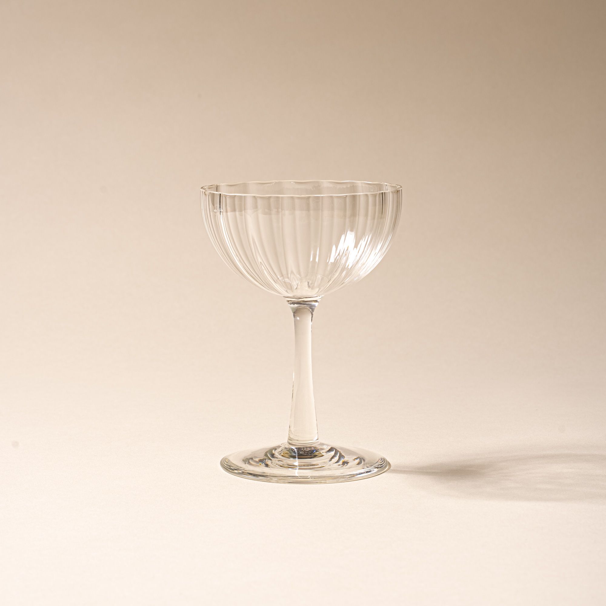 Clear ribbed glass coupe with a tall stem casting a soft shadow on a beige background.