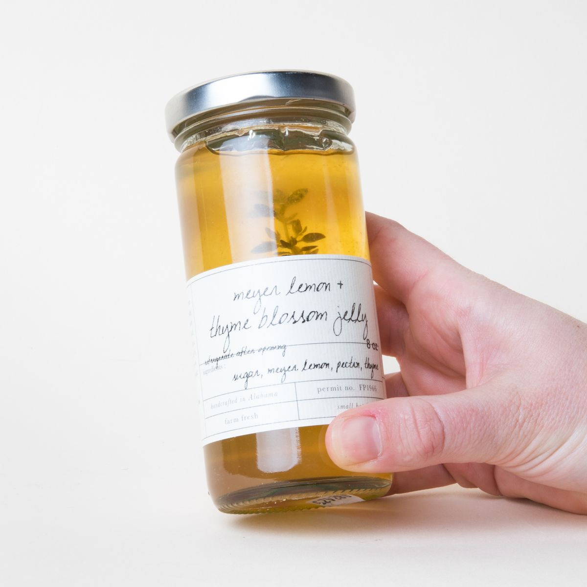 A hand holding a tall glass jar filled with yellow jelly with a white label that reads "Meyer Lemon and Thyme Blossom Jelly"