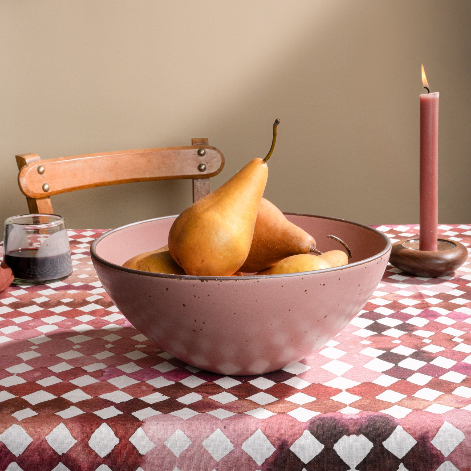 Large ceramic mixing bowl in a dusty mauve pink color filled with pears and citrus on a patterned tablecloth with a wooden chair in the background.