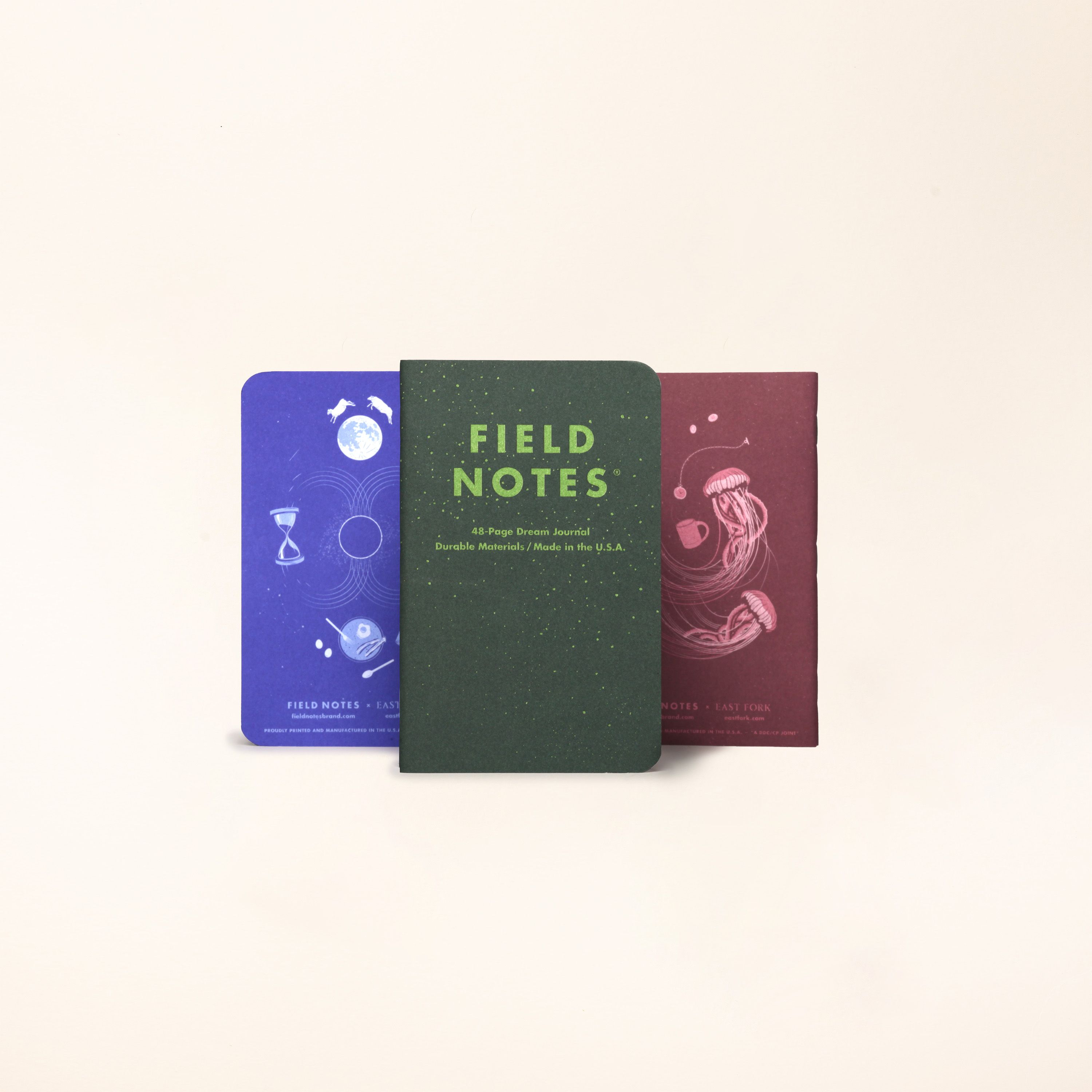 A 3 pack of Field Notes Journal standing up in a row with a forest green front facing one, and the left and right featuring their illustrated back covers in a lapis blue and plum color.