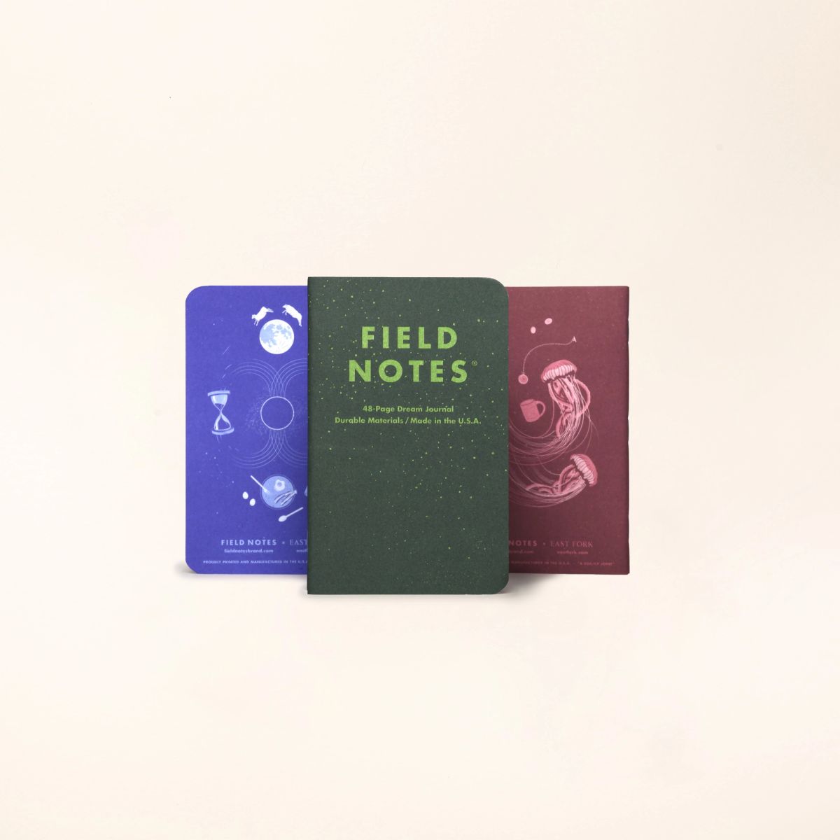 A 3 pack of Field Notes Journal standing up in a row with a forest green front facing one, and the left and right featuring their illustrated back covers in a lapis blue and plum color.