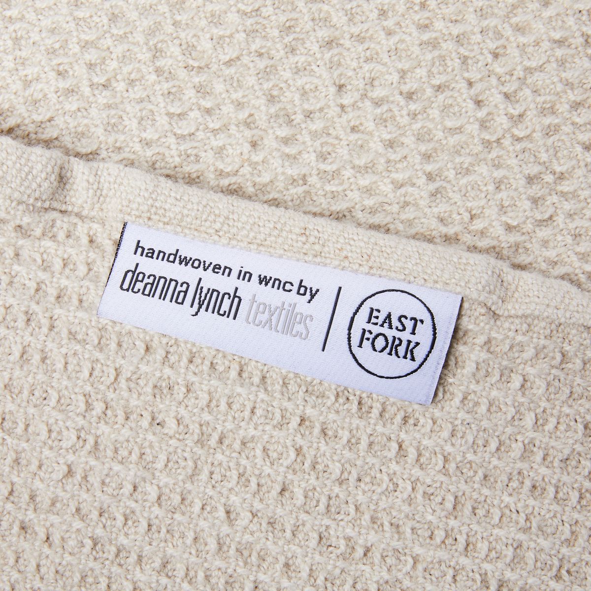 Close up of a natural color waffle towel with a white label that reads "Handwoven in WNC by Deanna Lynch Textiles" next to the East Fork logo
