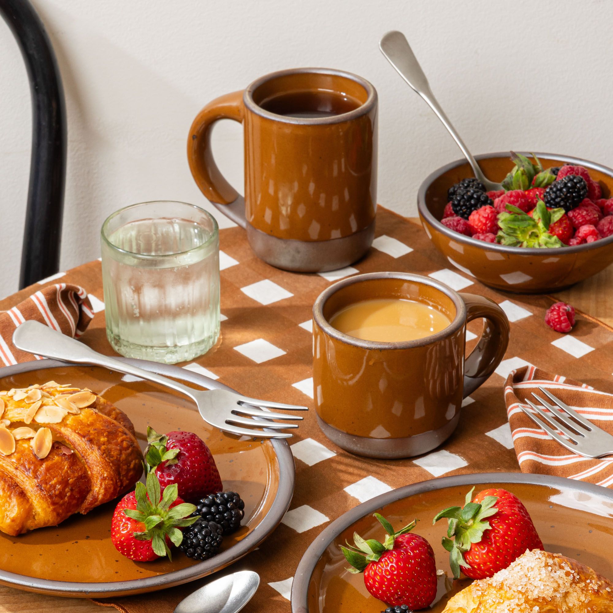Breakfast table with glossy caramel brown ceramic mugs, croissants, berries, and a bowl of raspberries on patterned linens.