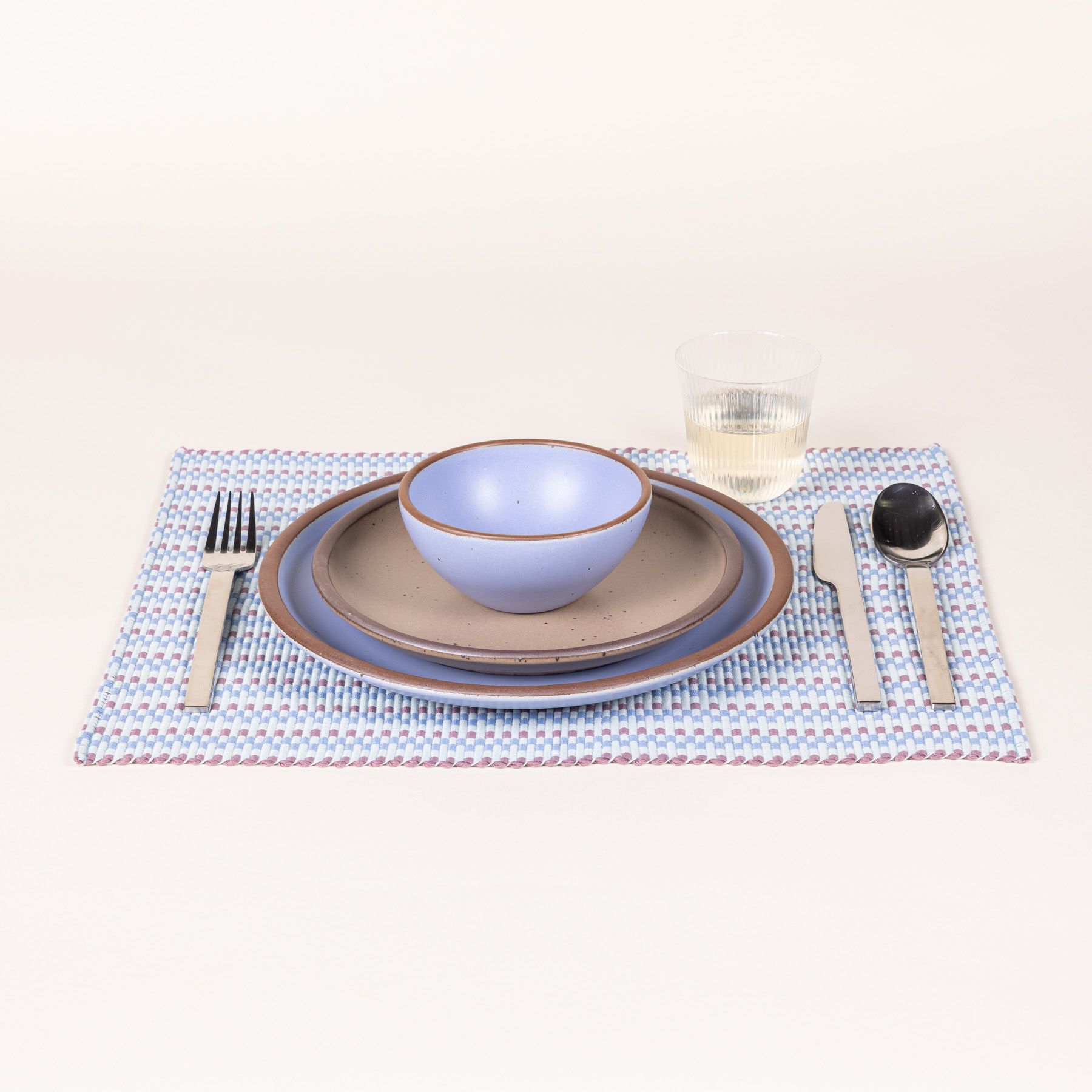 A place setting featuring a striped woven placemat with soft blue and purple colors, plates to match, a glass, and silverware.