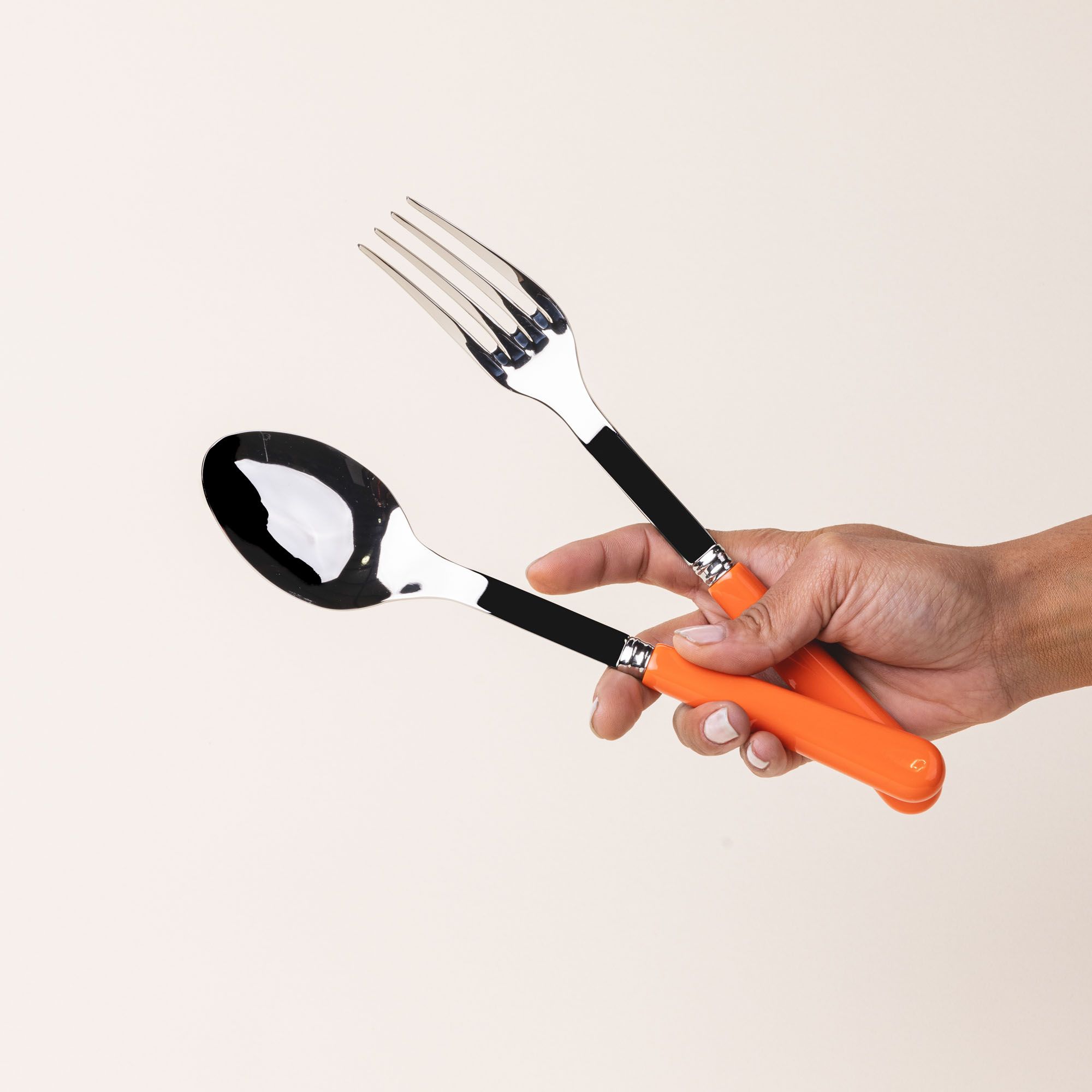 A hand holds a serving size spoon and fork in a shiny steel with a bold orange handle