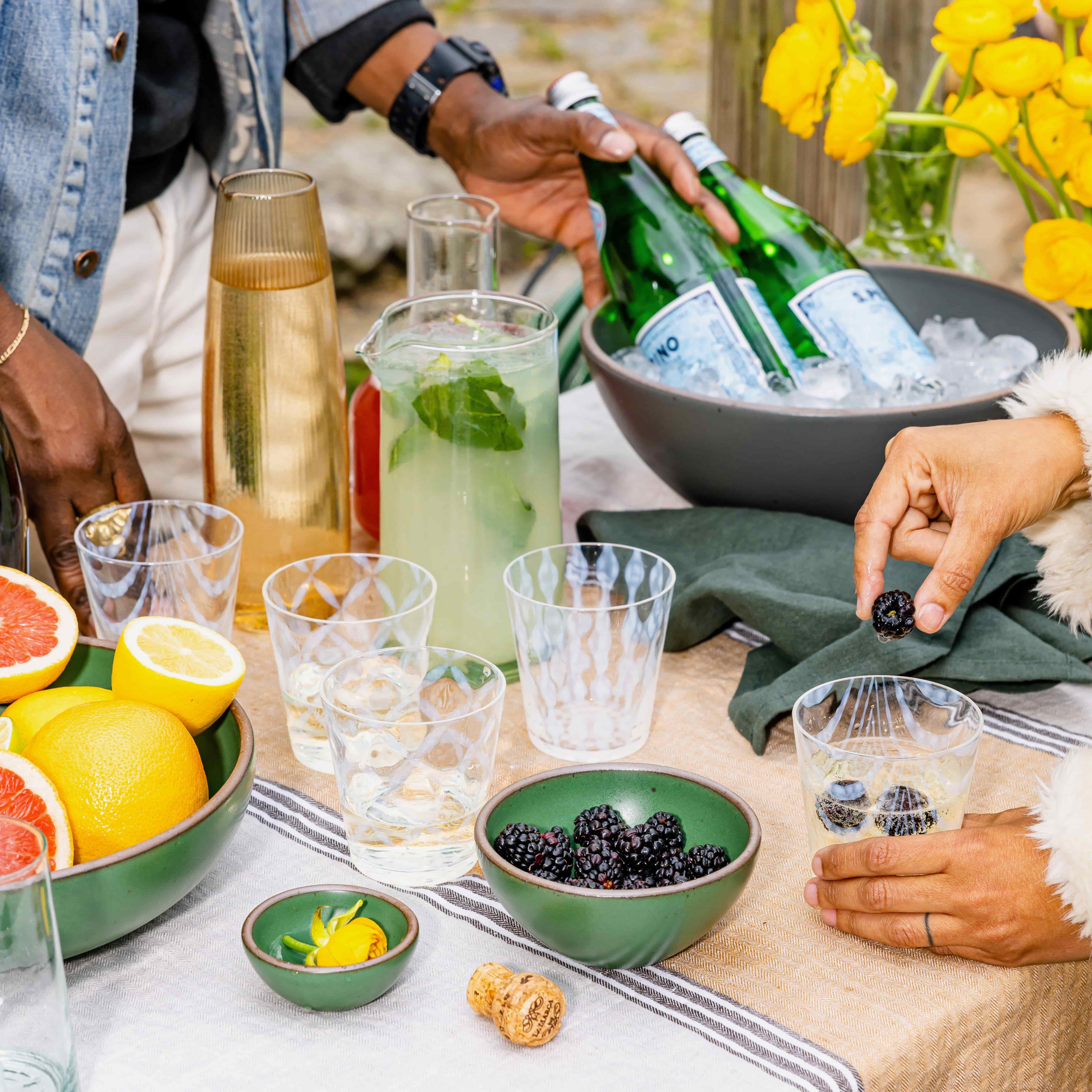 On a table outdoors is a little bar setup filled with various lowball glass tumblers, several glass pitchers, and fruits. A hand is dropping a blueberry in a drink and another hand is reaching for bottled seltzer.