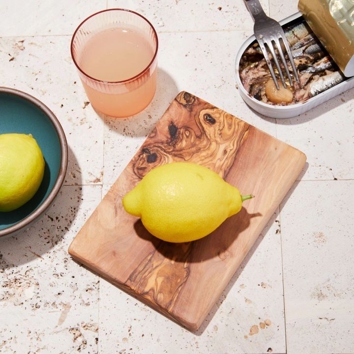 Square Olivewood Cutting Board with lemon on top, fish tin on the side, bowl with lemon and cup of lemonade