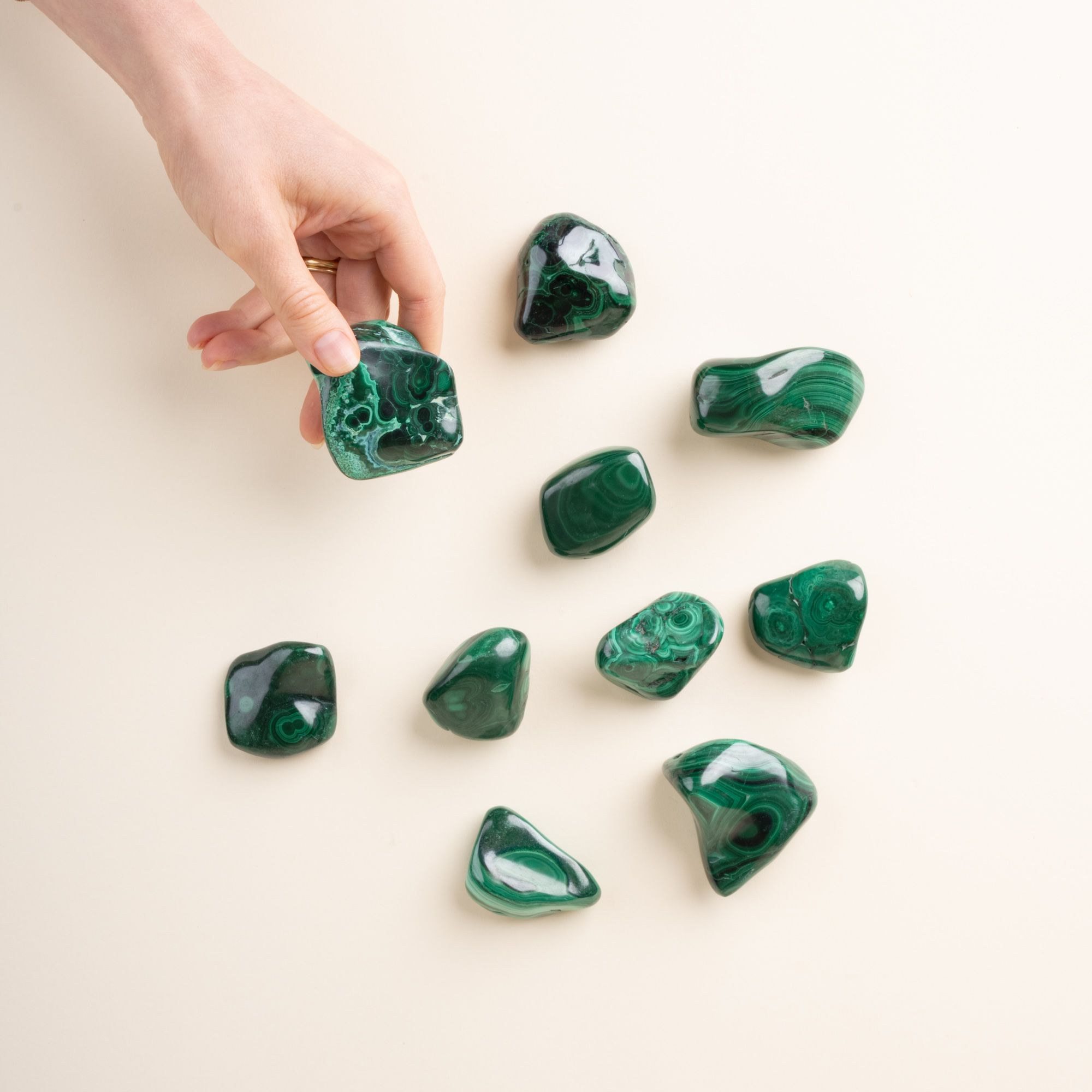 A hand holds one of a collection of polished green malachite stones with swirling patterns, scattered on a light neutral background.