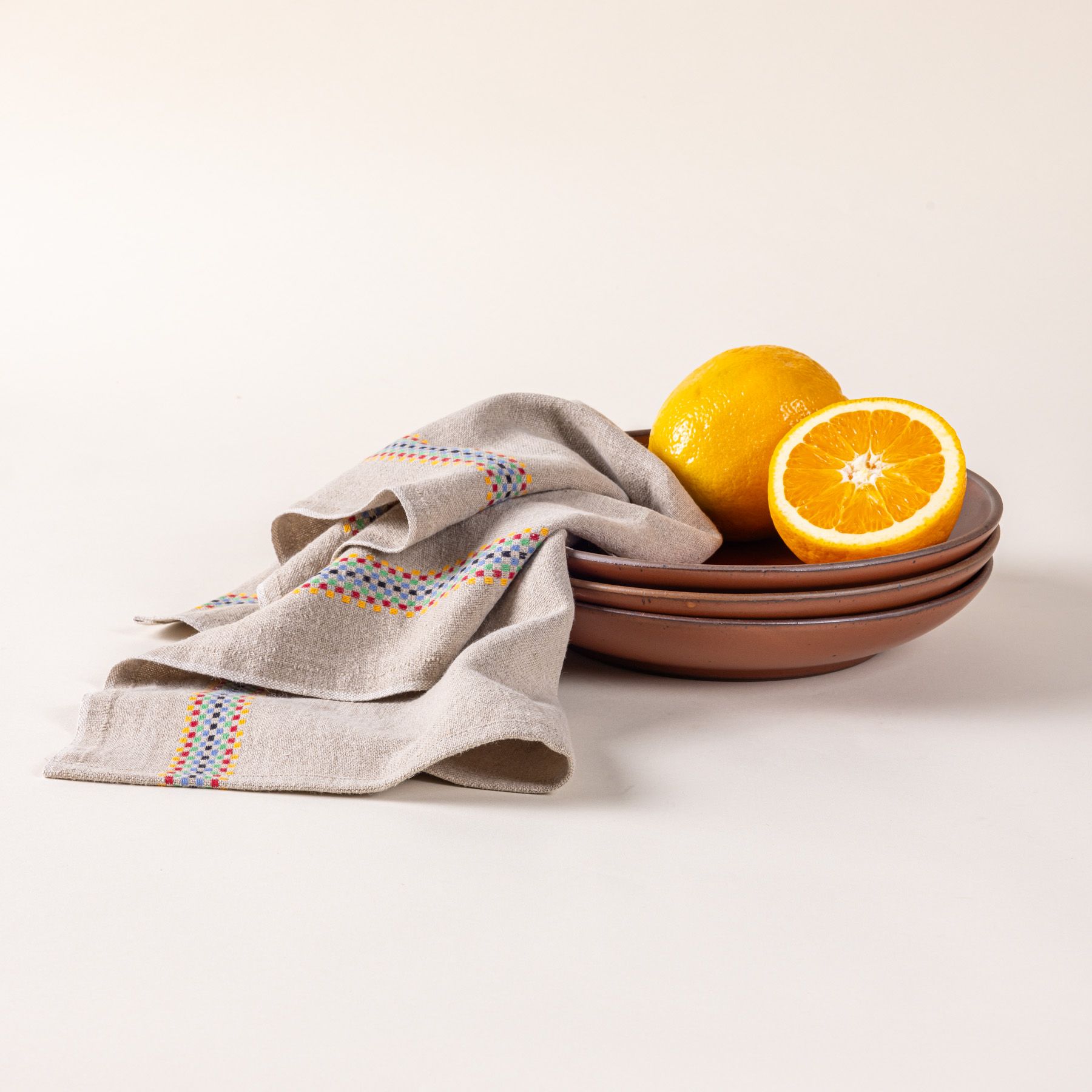 A linen towel in a beige color with a multicolor checkerboard stripe down the side with a stack of coupes and oranges