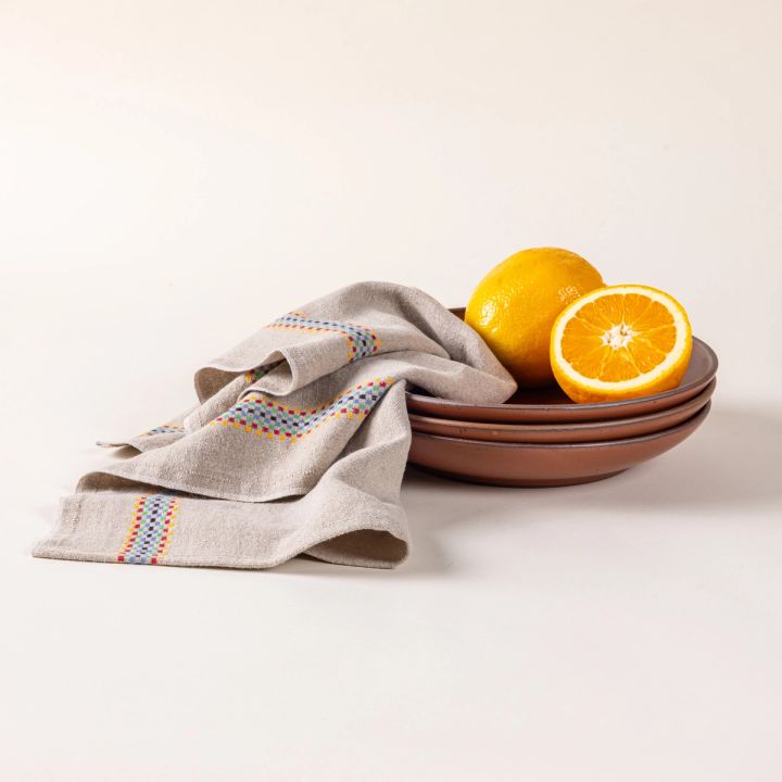 A folded linen towel in a beige color with a multicolor checkerboard stripe down the side with a stack of coupes and oranges