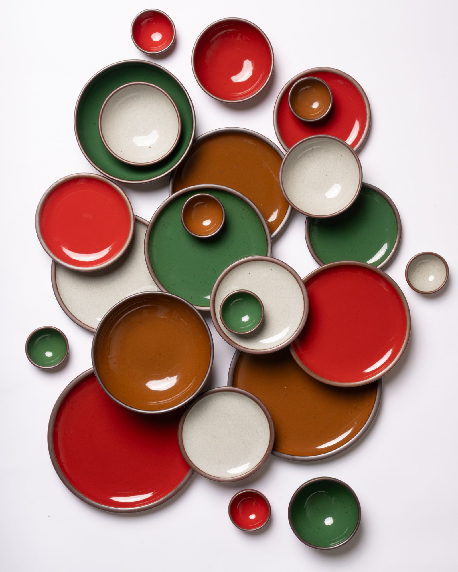An overhead of ceramic plates and bowls in a variety of sizes in glossy red, glossy green, glossy brown, and an icy reflective white color.