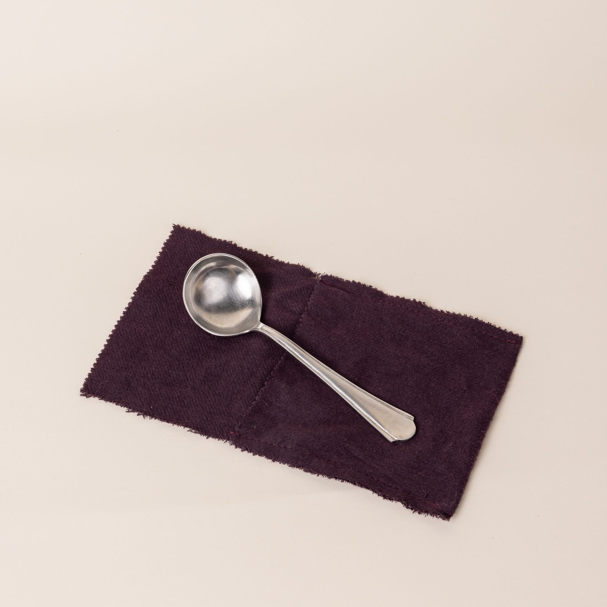 A silver spoon sits on a dark purple rectangle of fabric. The spoon has a skinny handle that gets a little wider and round on the end. The bowl of a spoon is circular.