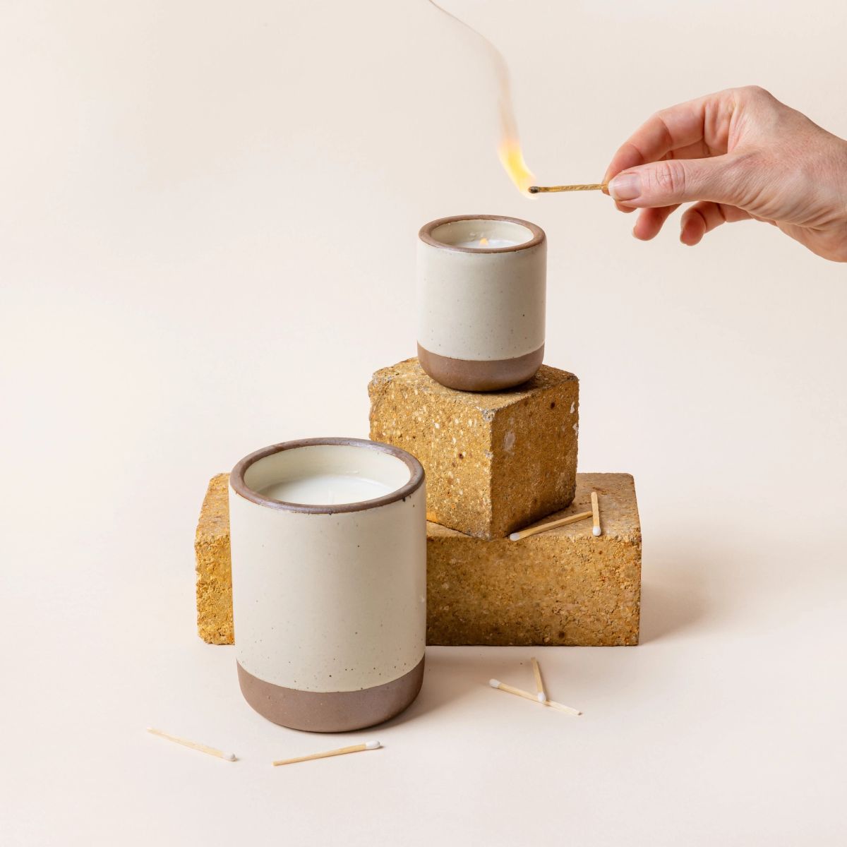 A large and small ceramic vessels in a warm, tan-toned, off-white color with candle inside each. A hand is holding a lit match to light the small candle. The candles are artfully arranged with brick and a few surrounding matches