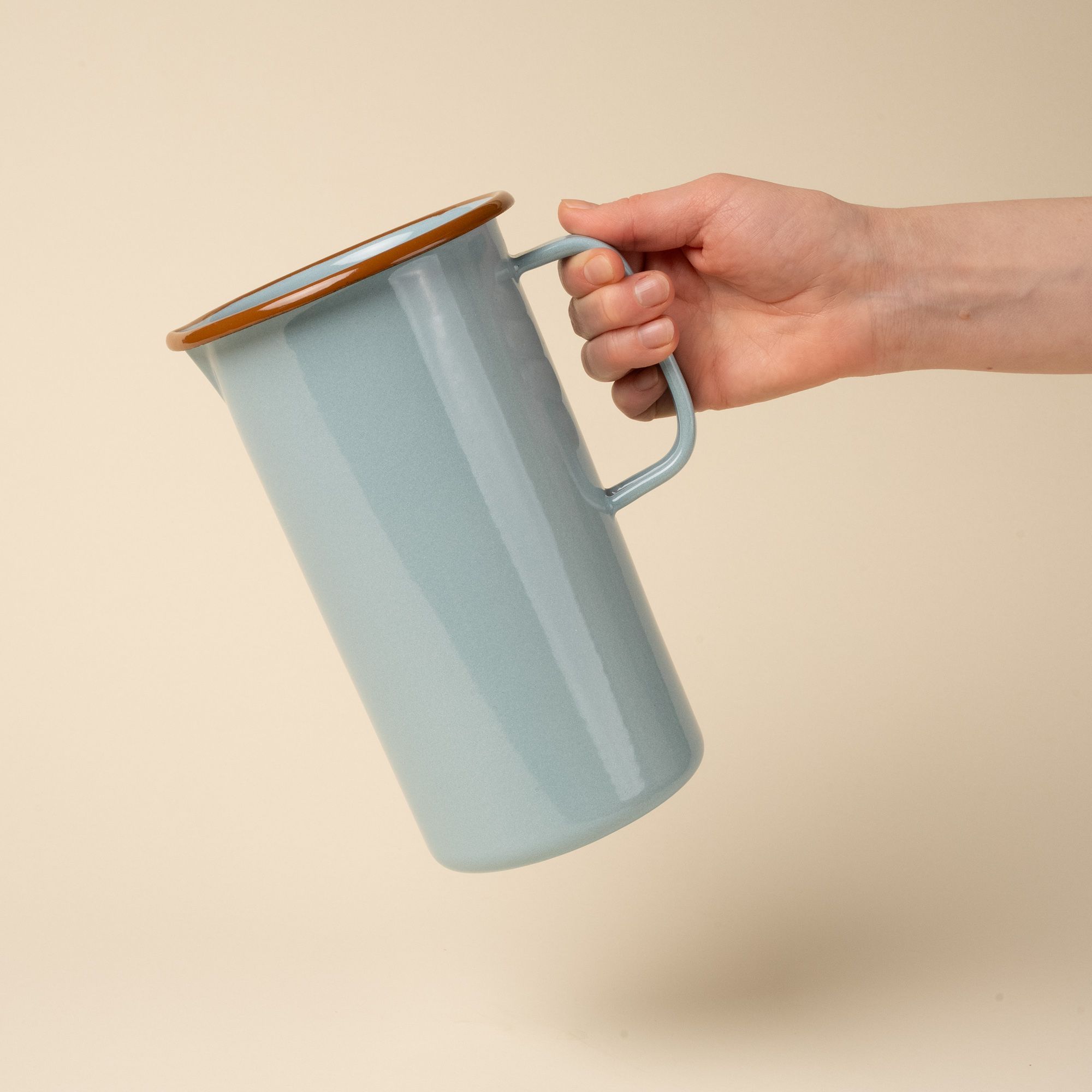 A hand holds a large enamel pitcher with handle in a robin's egg blue color with a warm brown rim.