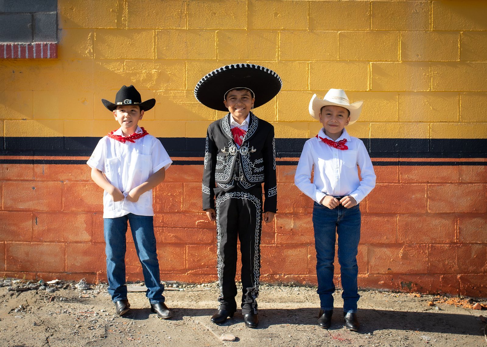 Three young boys are lined up in front of a building smiling at the camera. The boys on the left and right are wearing white shirts, jeans, and cowboy hats. The boy in the middle is wearing a traditional Mexican charro outfit in black.