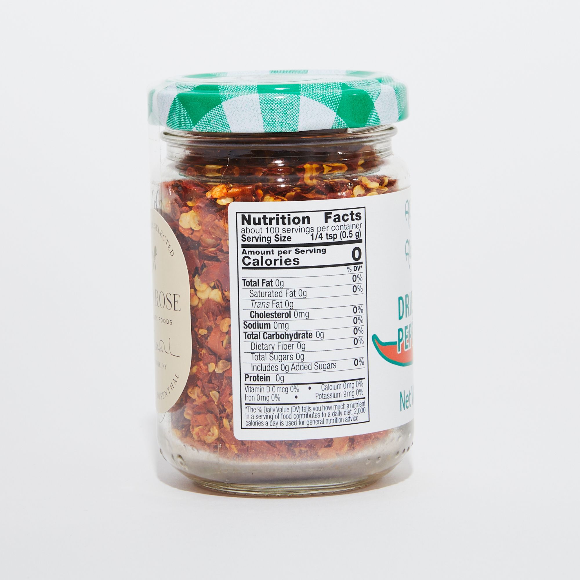 Clear glass jar full of red pepper flakes with a green and white gingham lid and a white label featuring nutritional facts