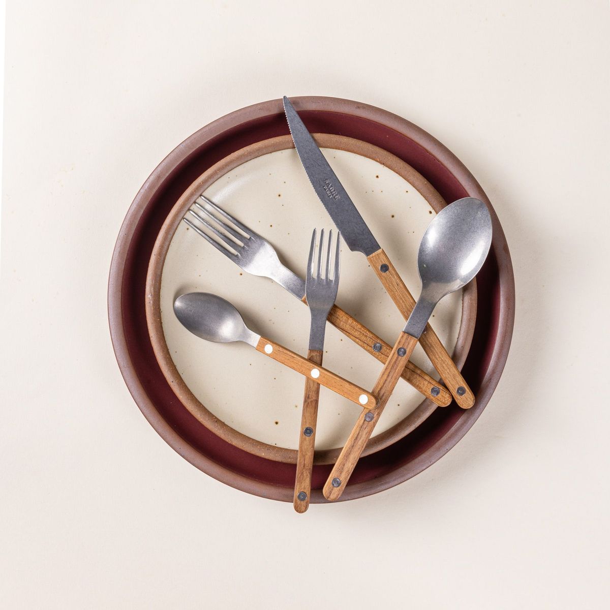 A set of 5 flatware pieces with a teak wood handle: a salad fork, dinner fork, knife, spoon, teaspoon sits on 2 ceramic plates