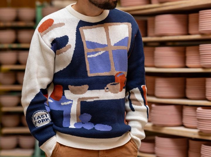 Alex wears a sweater with an illustration of a pottery workshop with a large window, shelves, and table with mugs and bowls. Alex stands in front of a rack of ceramic bowls in progress.