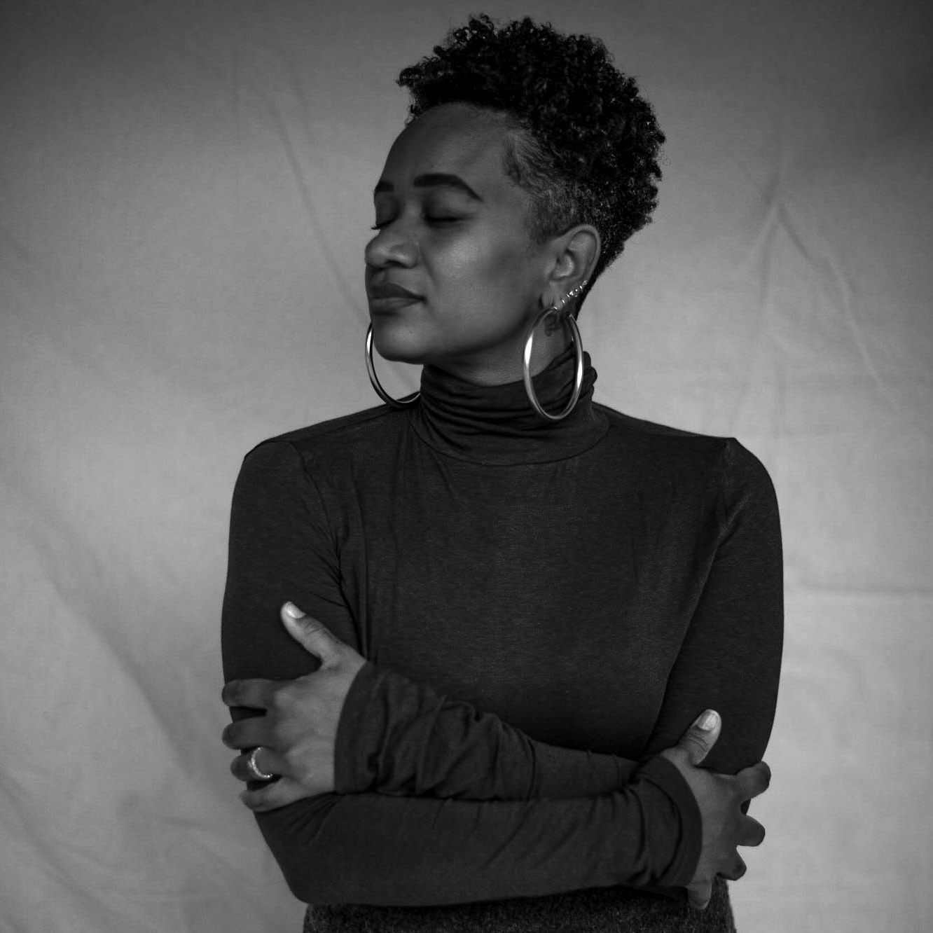 A black and white portrait of Alex Elle, a black woman wearing a dark turtleneck with her eyes closed and her arms crossed holding her.