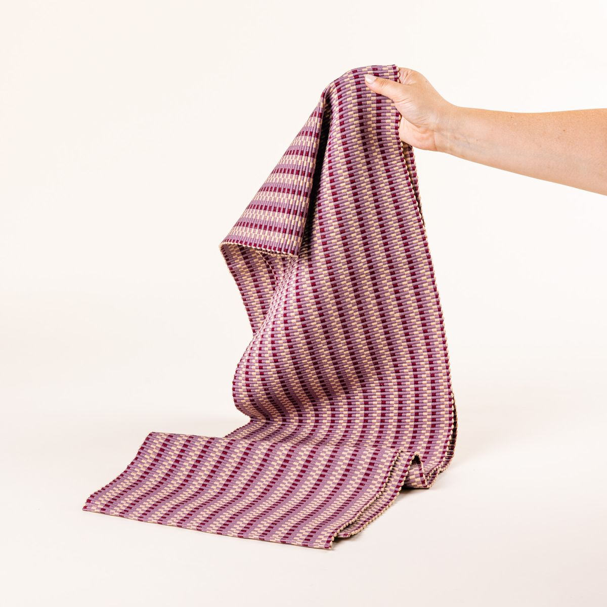 A hand holds up a woven table runner with thin horizontal striped tonal plum and blush colors