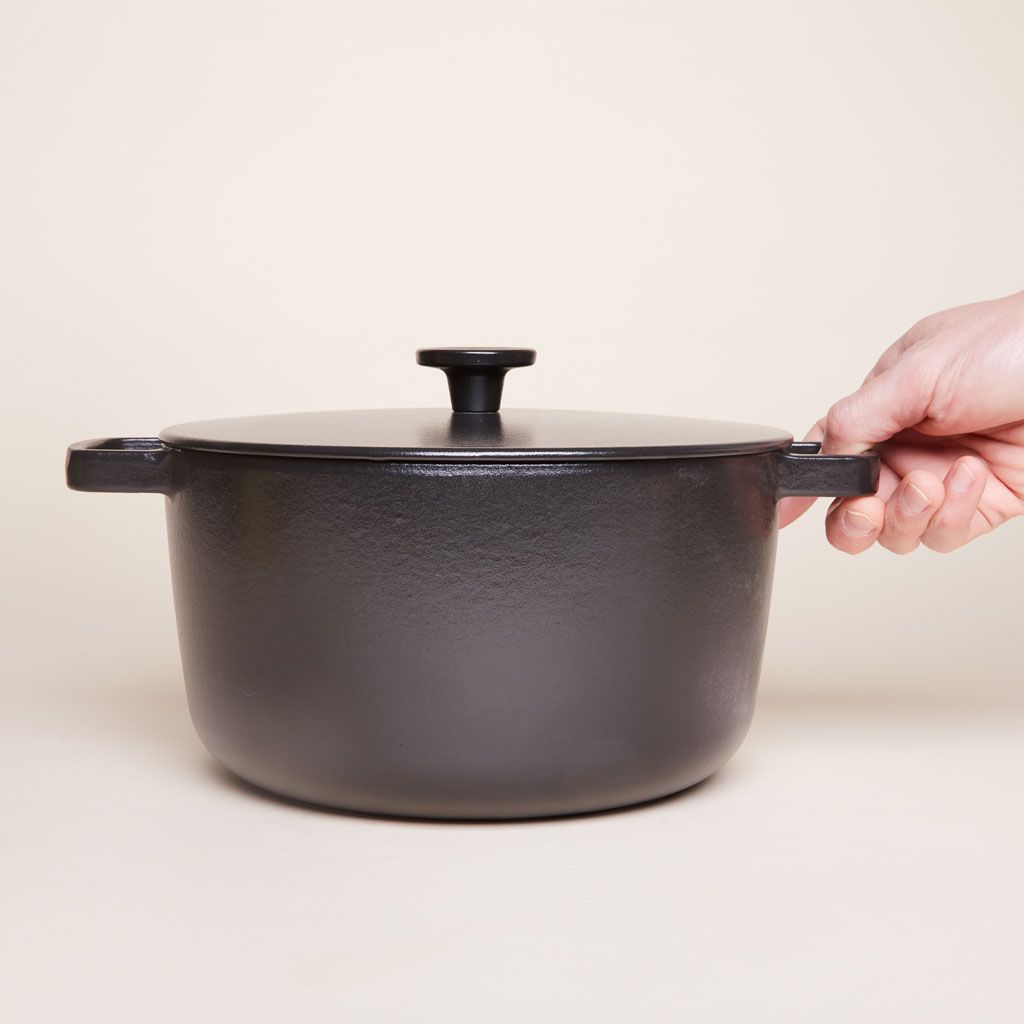 A hand touches the right handle of a tall black pot with two handles and a matching lid