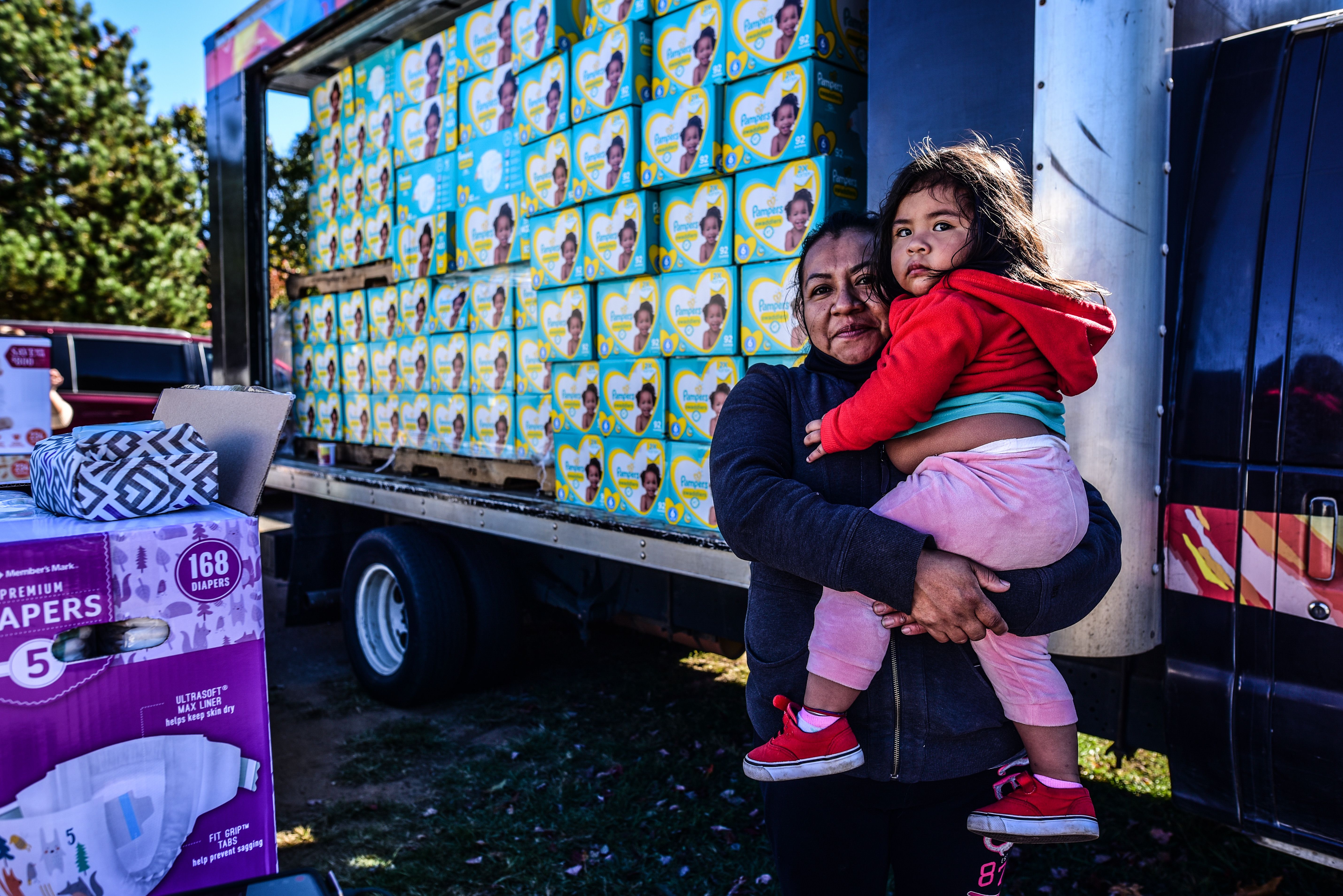 A person holds a child in front of a truck that is filled with boxes of diapers
