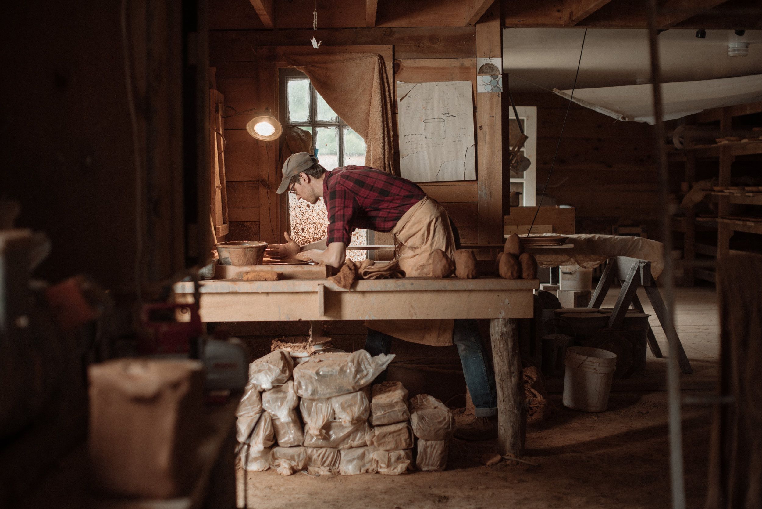 John working in a pottery studio, surrounded by formed clay, bags of clay, and tools.